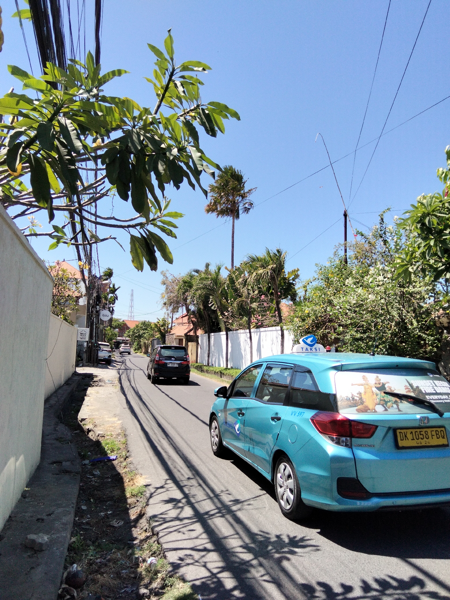 A bright, sunny day in Denpasar, Indonesia, with a clear blue sky. The image captures a residential street lined with a white wall on the left and lush greenery and houses with terracotta roofs on the right. A teal-colored taxi, with a "TAKSI" sign on its roof, is prominently featured in the foreground, driving away from the camera. The taxi has a colorful decal on its rear window depicting figures in traditional attire, and a license plate that reads "DK 1058 FBQ". Other vehicles, including a black SUV, are also visible on the road. Overhead, a tangle of electrical wires stretches across the sky, casting shadows on the street. The presence of palm trees and tropical foliage suggests a warm climate. The overall scene depicts a typical day on a street in Denpasar.