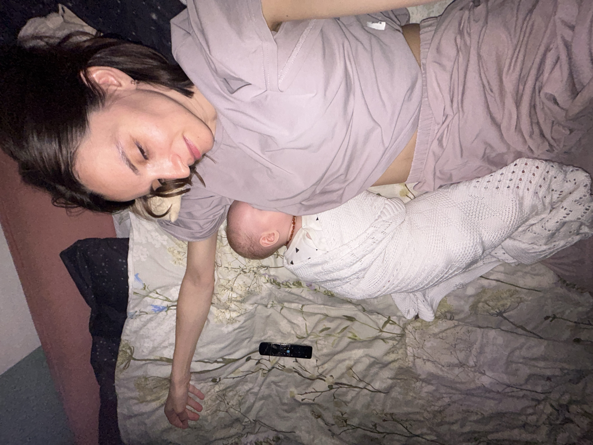 A woman with short brown hair lies on a bed next to a baby wrapped in a white blanket. The woman is wearing a pink t-shirt and matching pants. She is lying on her back with her eyes closed and her head turned to the left. The baby is lying on its side next to her chest. A black TV remote control is on the bed near the woman's hand. The bed has a floral patterned sheet and is near a pink wall. The location is unknown and cannot be identified from the image.