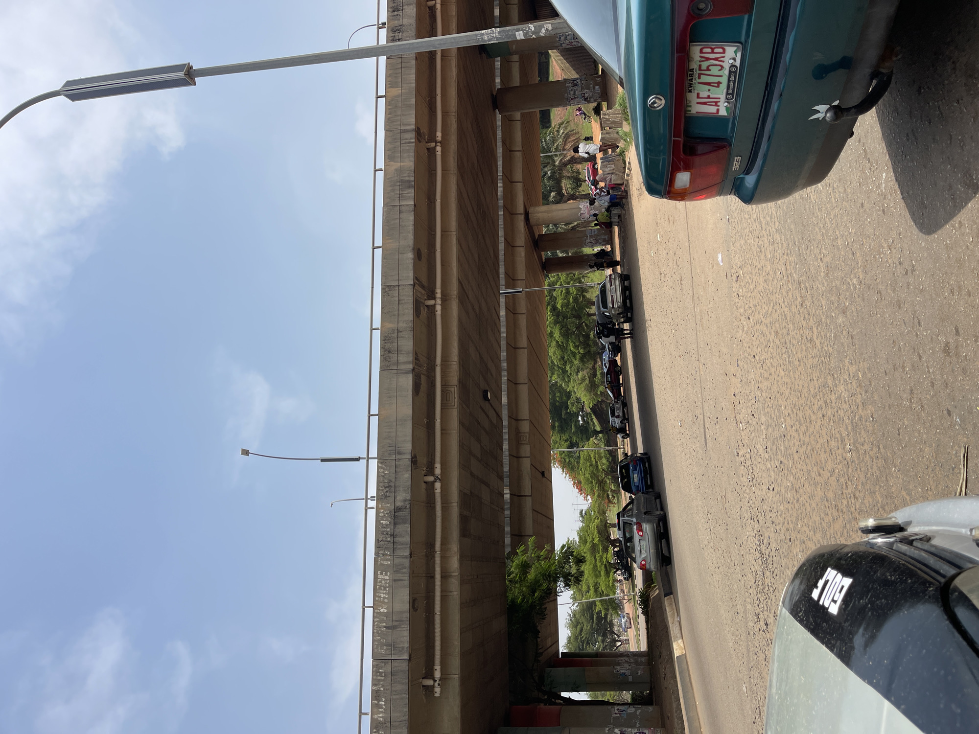 The image depicts a street scene in Abuja, Nigeria, under a concrete overpass. The sky is visible on the left, featuring light clouds. The overpass is constructed of stacked concrete blocks. Streetlights line the side of the overpass.
The street is lined with parked cars. A green Mazda 323, with the license plate “LAF-475XB” (KWARA), is visible in the upper right of the image, and a gray Volkswagen Golf partially appears in the lower right. A line of other vehicles are parked near the center of the image.
People are gathered under the overpass, in the upper part of the image, but are not clearly visible. The road surface is worn.