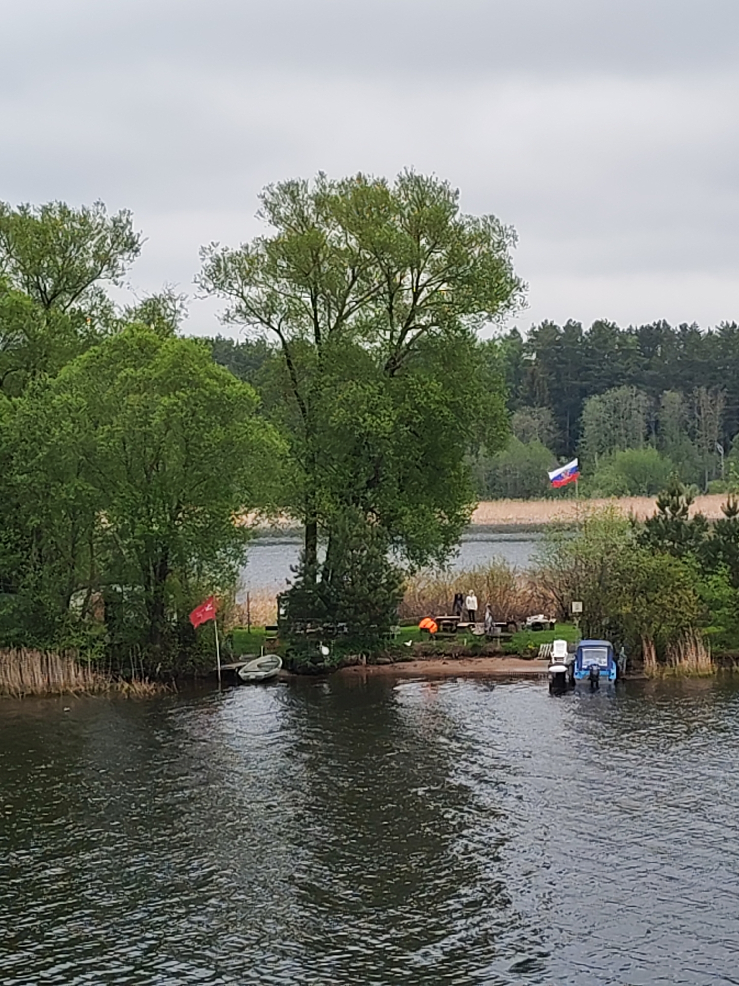 The image depicts a view of a lake or river with a small island or peninsula in the middle. The island has several trees, a small dock, and two boats. There are also several people standing on the island near a table. Two flags are visible; one red flag is on the left of the island near a small white boat, and a Russian flag is seen in the background of the island. The water is calm with some ripples. The sky is overcast. The image is reported to be taken in Novomelkovo, Russia.