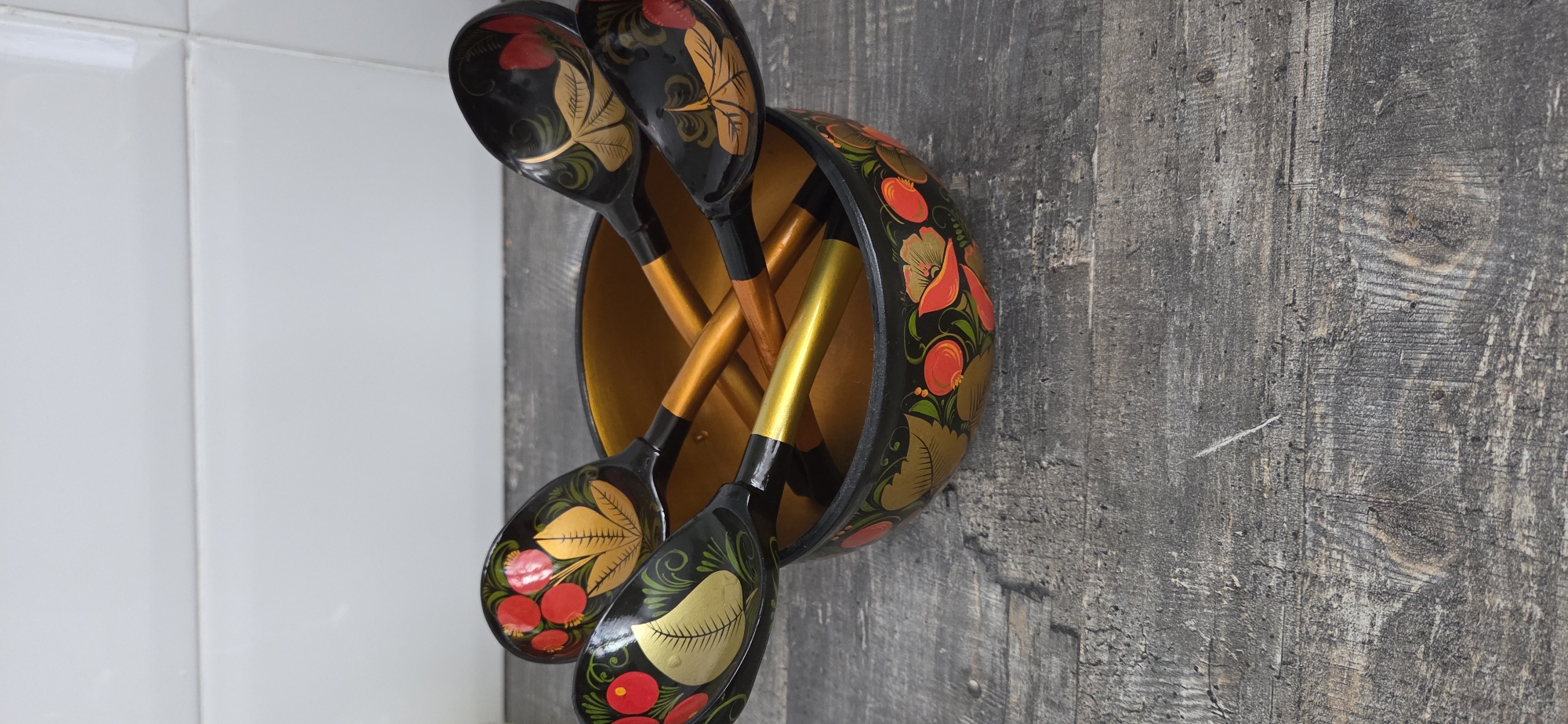 This image captures a close-up of a set of traditional Russian Khokhloma-style kitchenware. The primary subjects are a decorative wooden bowl and several wooden spoons, all intricately hand-painted.
The bowl is black on the exterior, adorned with vibrant floral and berry patterns in shades of red, gold, and green. The interior of the bowl is a bright golden color. Inside the bowl, several matching wooden spoons are stored. These spoons also feature black heads with similar red, gold, and green floral and berry motifs. Their handles are a contrasting golden color, complementing the bowl's interior.
The items are arranged against a two-part background. To the left, a clean, white tiled wall with visible grout lines suggests a kitchen or bathroom setting, though the latter is less likely given the objects. To the right, a dark, textured wooden wall or paneling provides a rustic contrast. The wood shows visible grain, knots, and a weathered appearance.
There is no specific activity taking place other than the display or storage of these items. No people are visible, and no text is present in the image. The lighting suggests an indoor setting, but the time of day or weather cannot be determined. The image itself appears to be rotated 90 degrees clockwise from its original orientation. The overall impression is one of traditional craftsmanship and decorative art.
