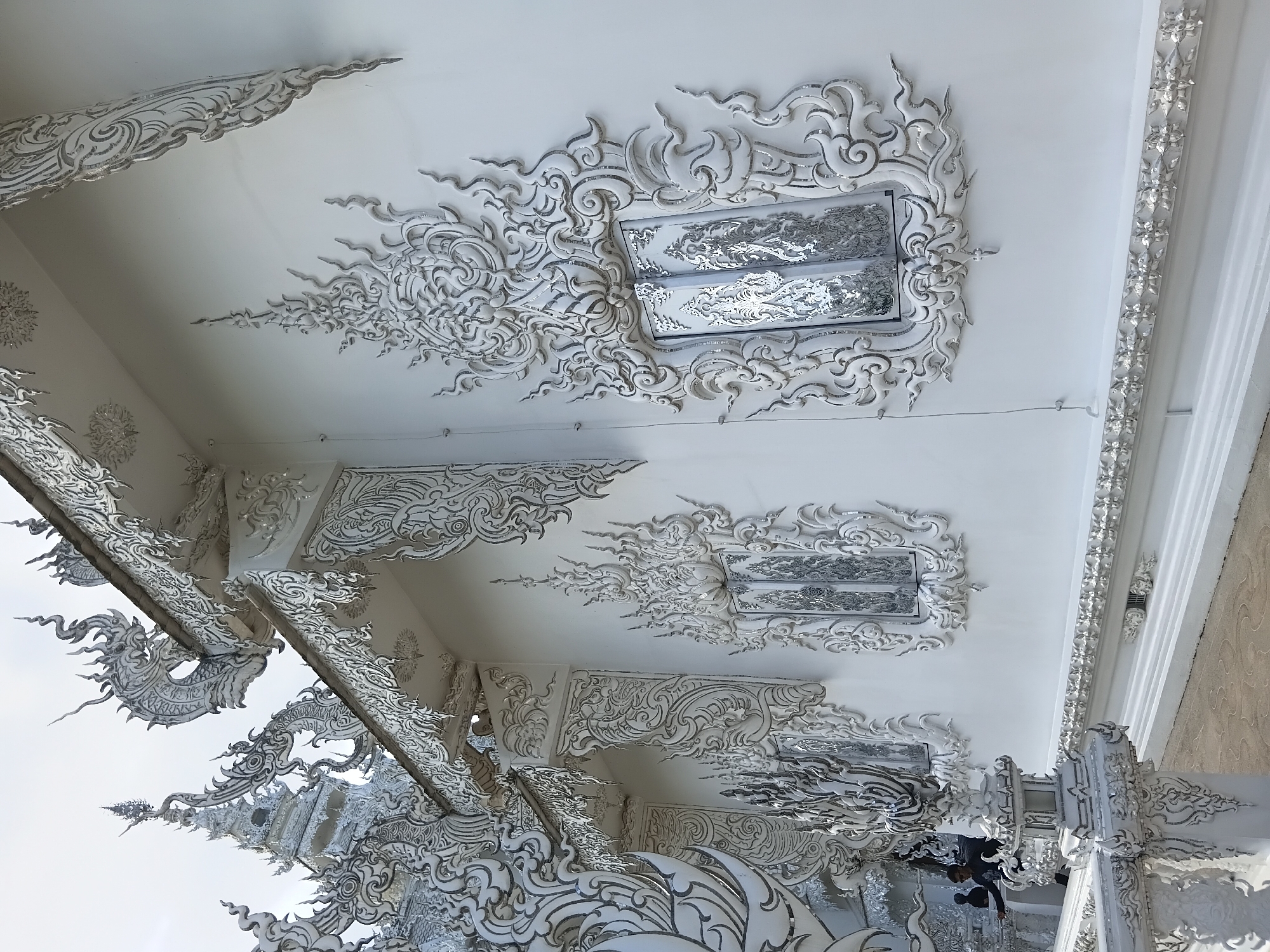 The image shows a detailed exterior of a white temple building in Ban Rong Khun, Thailand. The architecture is characterized by intricate white stucco ornamentation and decorative elements resembling mythological creatures, particularly dragons, along the roofline and building walls. Several windows are visible, featuring ornate frames and mirrored or reflective panels. The building's surface is pristine white, contrasting with the gray sky. In the lower left of the frame, a person in dark clothing and sunglasses is visible, interacting with the structure. The ground in front of the building appears to be paved. The overall scene depicts the elaborate and distinctive design of the White Temple.
