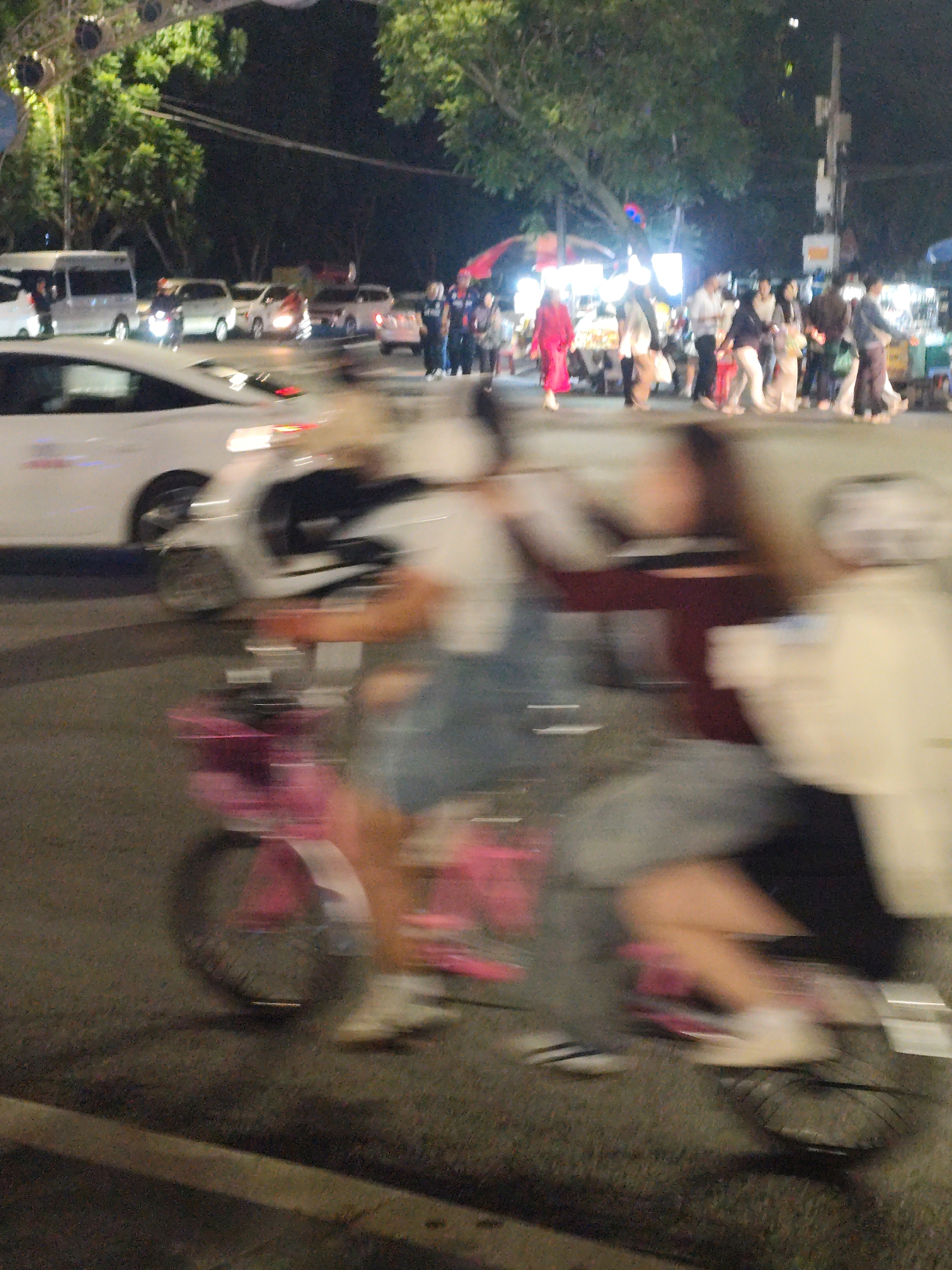 The image shows a street scene at night in what appears to be Da Lat, Vietnam. A group of people is riding bicycles across the street in the foreground, creating motion blur. In the background, pedestrians are walking on a sidewalk near what appears to be market stalls or shops. There are several vehicles parked along the street, including vans and cars. The scene is illuminated by streetlights and lights from the stalls.