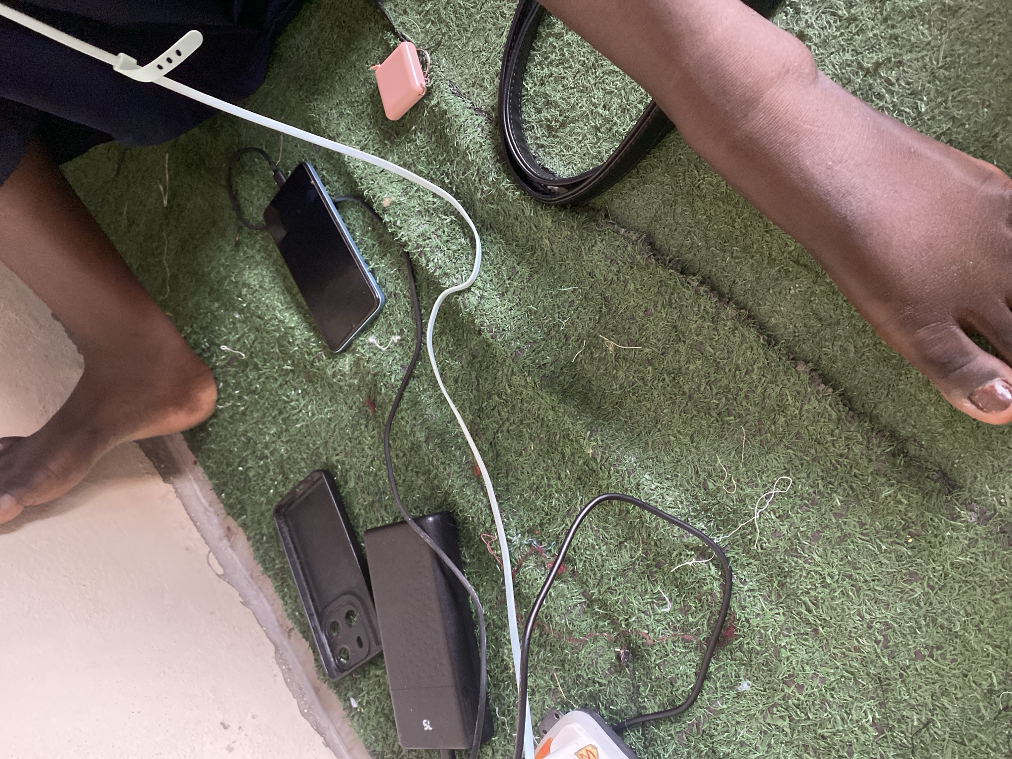 The image shows a close-up view of a person's legs and feet resting on artificial turf, possibly indoors. Several electronic devices and accessories are scattered on the turf, including a black smartphone plugged into a charger, a black phone case, a black power bank, and a small pink square object that appears to be a portable charger or case for earbuds. A black belt is also visible. The lighting suggests an indoor environment with artificial turf flooring. The scene is located in Jauro Yinu, Nigeria.