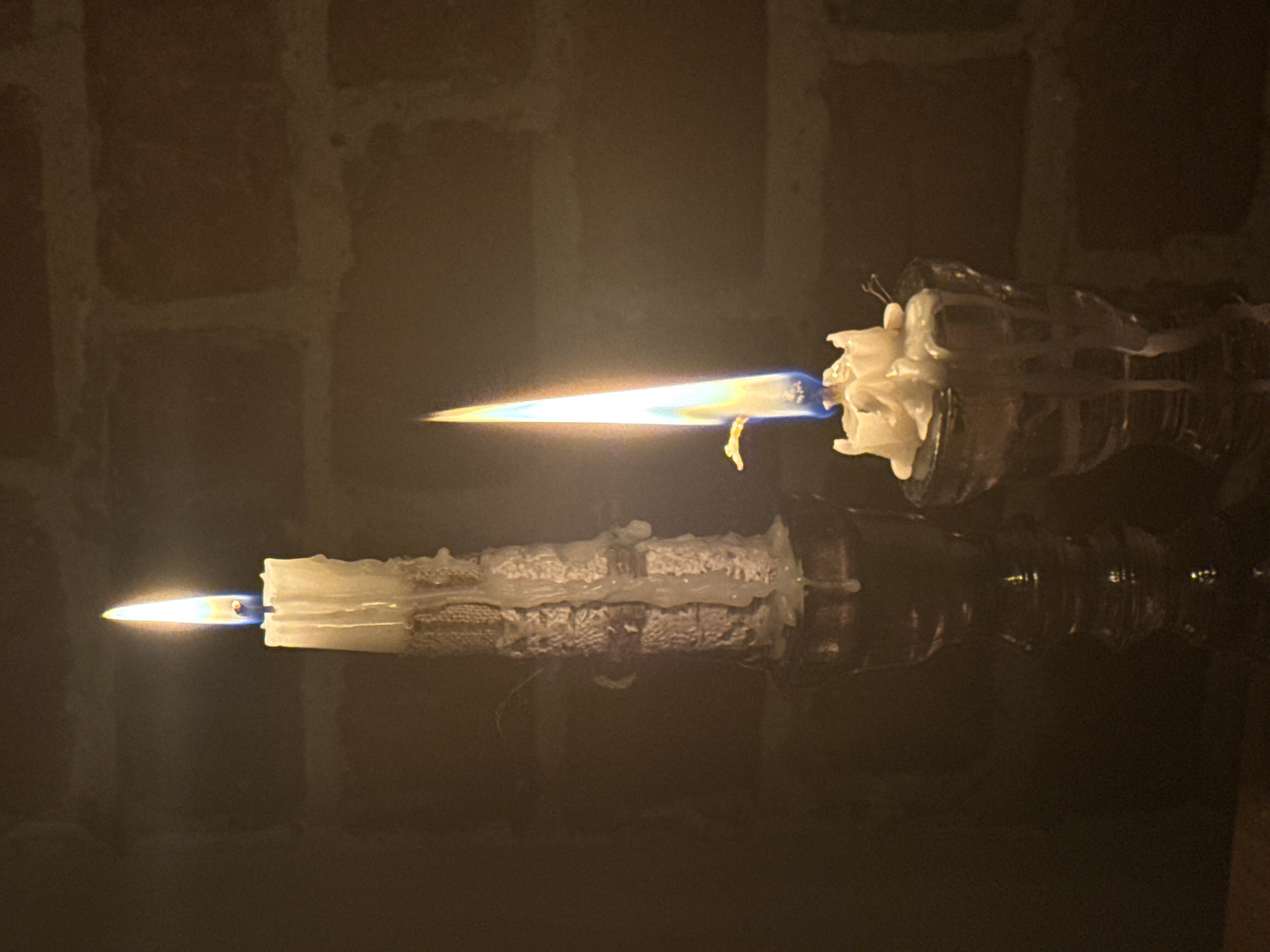 This media file captures two white candles burning in a dimly lit, indoor setting. Both candles are oriented horizontally, with their flames pointing towards the left against a dark, reddish-brown brick wall backdrop.
The upper candle exhibits a striking, unusually long and sharp flame, extending significantly from the wick. This flame displays vibrant orange and yellow hues, with a distinct blue base closer to the candle. Abundant melted wax drips down the sides of this candle, forming intricate patterns and textures. Below it, a second white candle also burns, but with a more conventional, shorter, and teardrop-shaped flame, primarily glowing in warm orange-yellow tones.
Both candles and their flames are clearly reflected on a dark, possibly polished or glassy surface beneath them, indicating placement on a reflective table or stand. The scene is illuminated almost entirely by the candle light, suggesting a dark room or nighttime. No people are visible, and there is no discernible text. The image provides a sense of quiet ambiance, with the long flame on the top candle being a notable and unusual detail, possibly caused by a draft. The location context provided is Rostov-on-Don, Russia.
