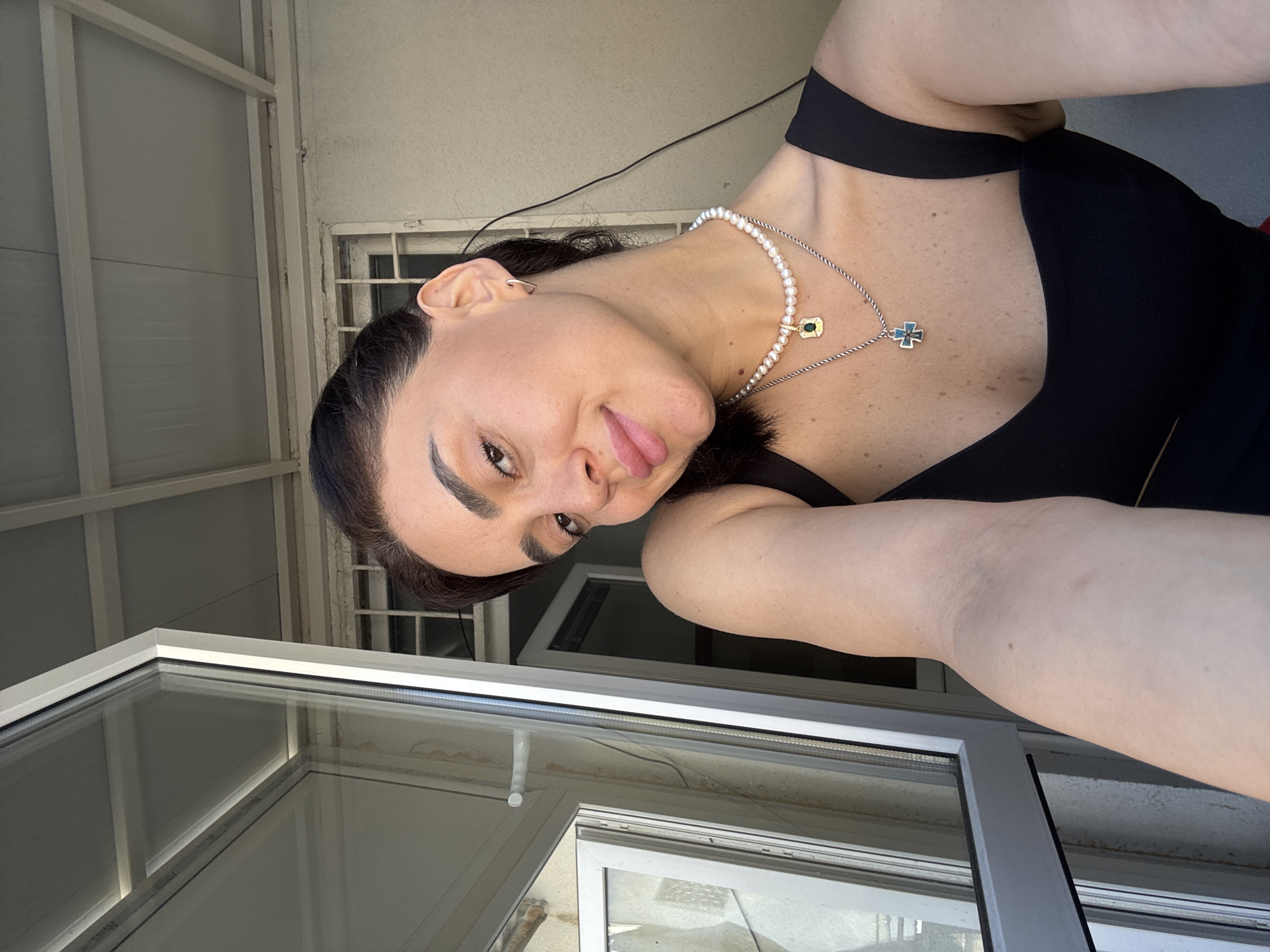 A woman with dark hair and fair skin, wearing a black tank top and necklaces, takes a selfie in front of an open window. The background shows architectural details characteristic of Tbilisi, Georgia, including a weathered building facade with barred windows and a corrugated metal awning. Sunlight illuminates the scene.