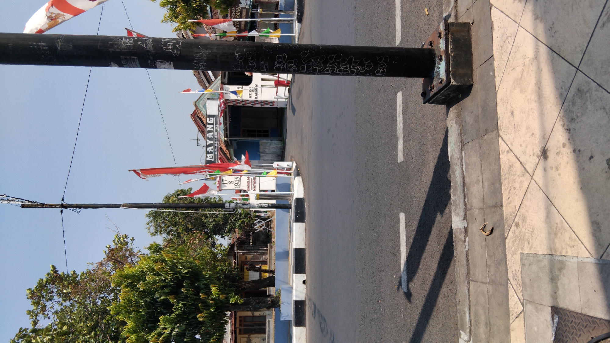 The image captures a street scene in Batang, Indonesia, likely in the vicinity of the "Mayang" area, as indicated by signage. A dark, thick pole, possibly a utility pole, dominates the right side of the frame, exhibiting graffiti. A street intersection is visible with a paved road and a sidewalk. Several Indonesian flags, red and white, are displayed, some mounted on poles and others attached to buildings, suggesting a festive or patriotic occasion. One prominent sign reads "MAYANG" with accompanying text "RT. 04 RW. 02". Another sign on a white post displays "17 8 1945", referencing Indonesian Independence Day. Buildings line the background, appearing to be simple structures. Trees with green foliage are present on the left side. The sky is clear and blue. The overall scene suggests a public space, possibly in a residential or commercial district of Batang.