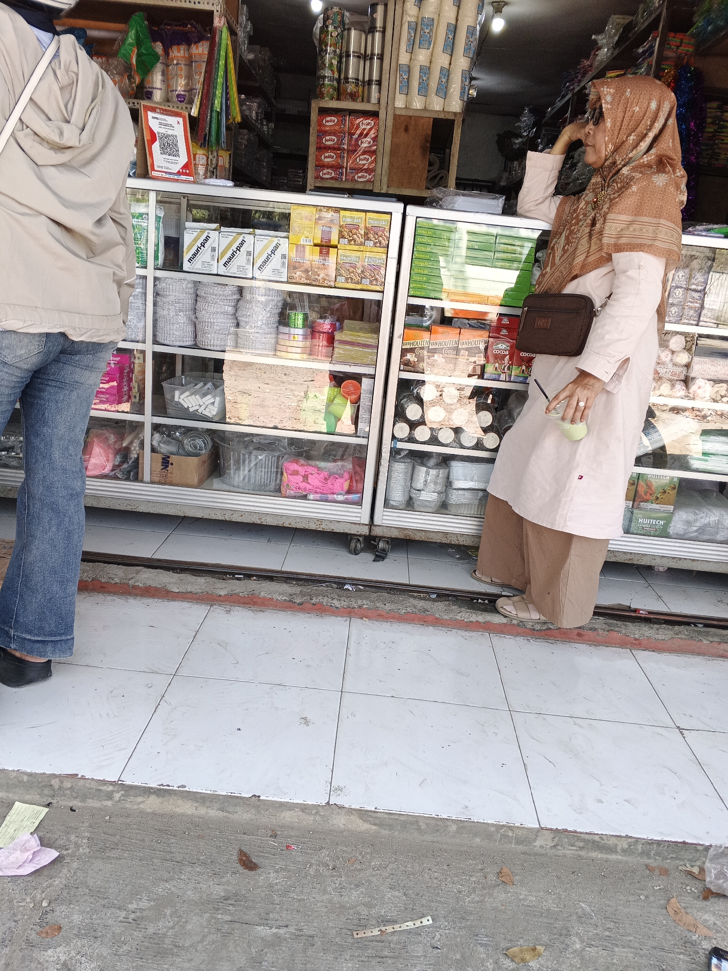 The image shows a street-level view of a small shop in Sukabumi, Indonesia. A woman wearing a beige headscarf and a light pink outfit is standing in front of the shop, holding a green drink. She is looking to her left. Another person, only their lower body is visible, is standing to the left of the woman, wearing blue jeans and a light beige jacket. The shop appears to sell various goods, with shelves filled with packaged items, including food products, disposable containers, and other household items. The shop's facade is a display case with glass doors. The ground in front of the shop is tiled.