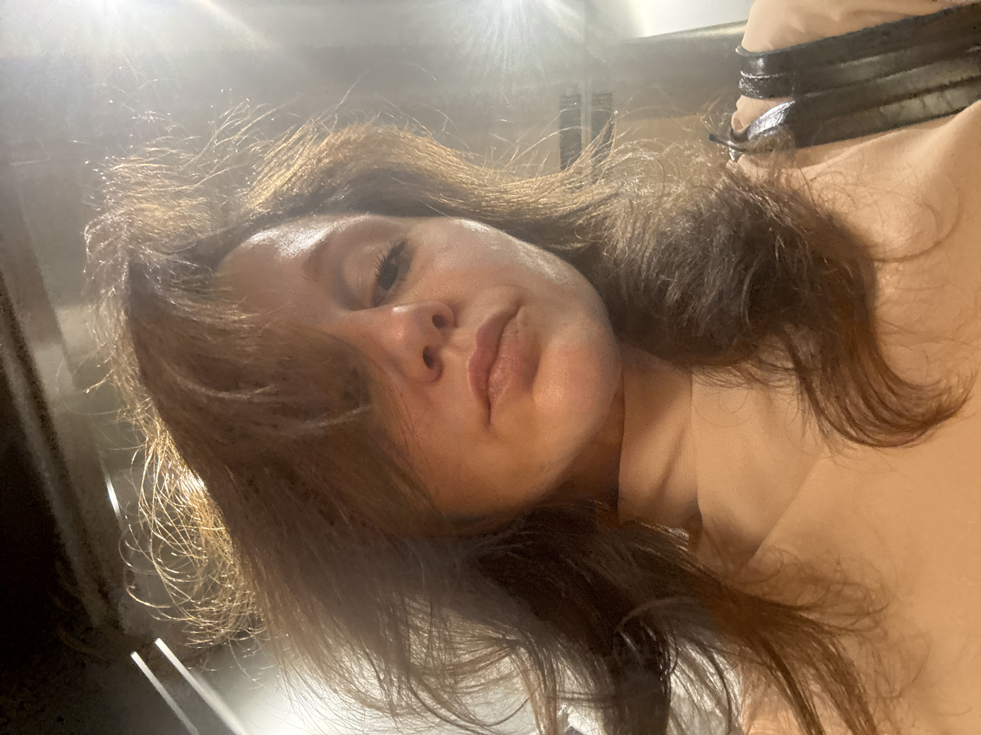 A selfie of a woman with brown hair, light skin and a neutral expression, taken at an upward angle with bright light shining from above. The location is unidentifiable as the background is out of focus. There is no evidence to suggest the location is in Dubai, United Arab Emirates.