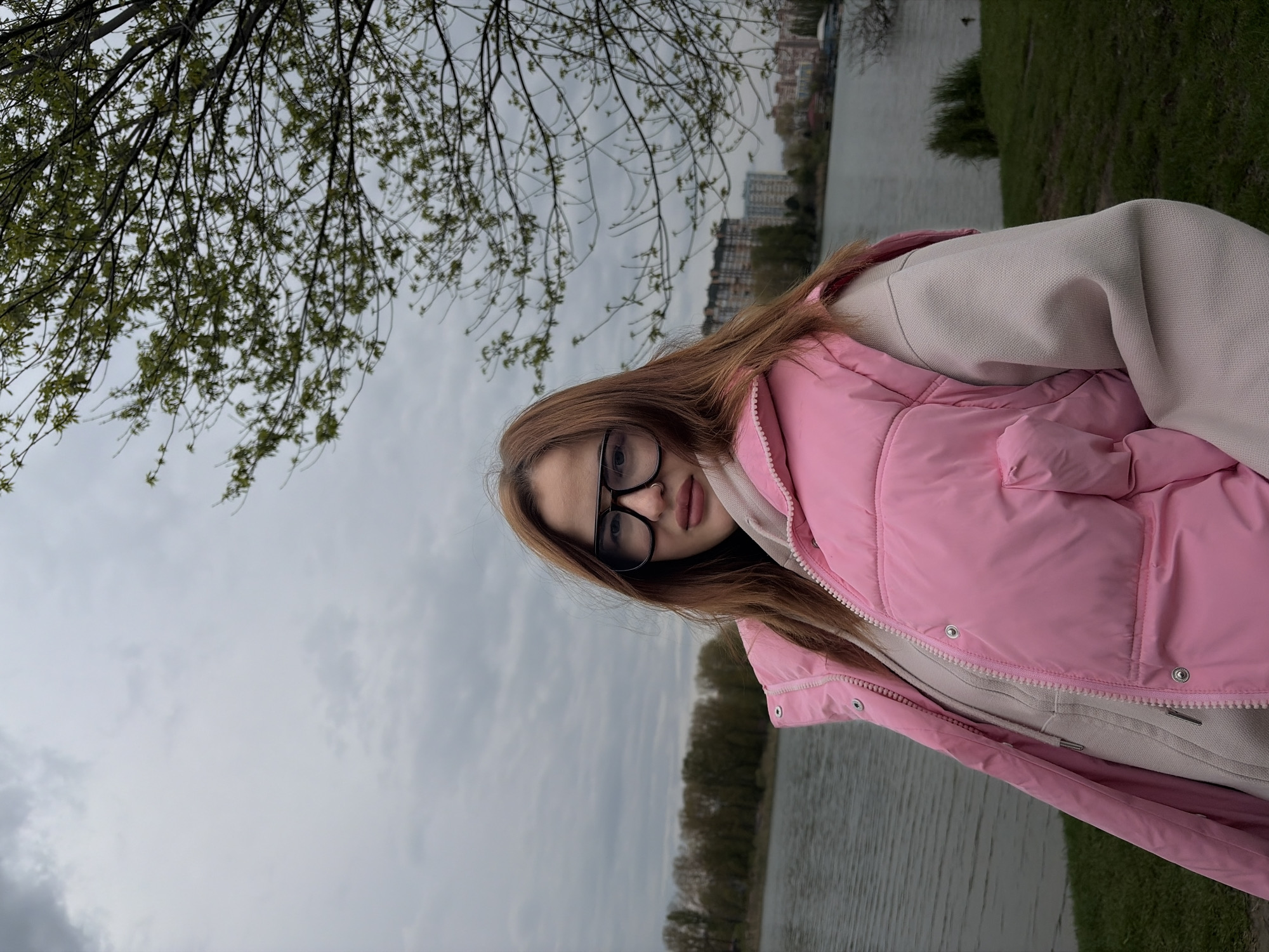 A young woman with long, straight, reddish-brown hair is depicted in a portrait-style photograph, taken outdoors. She wears black-rimmed glasses and a subtle nose piercing. Her attire consists of a light-colored (possibly beige or cream) hooded sweatshirt, layered under a vibrant pink puffer vest. Her expression is neutral as she looks towards the camera.
The scene is set alongside a body of water, likely a river or lake, with a grassy bank visible to the right. In the background, beyond the water, there are trees with sparse, light green foliage, indicating springtime. Further in the distance, an urban skyline with multi-story buildings can be seen. The sky is heavily overcast and grey, suggesting a cloudy day. Overhanging tree branches with budding leaves frame the upper left portion of the image. The overall impression is a casual outdoor moment. This scene is located in Krasnodar, Russia, likely along a park or river embankment. There is no visible text in the image.