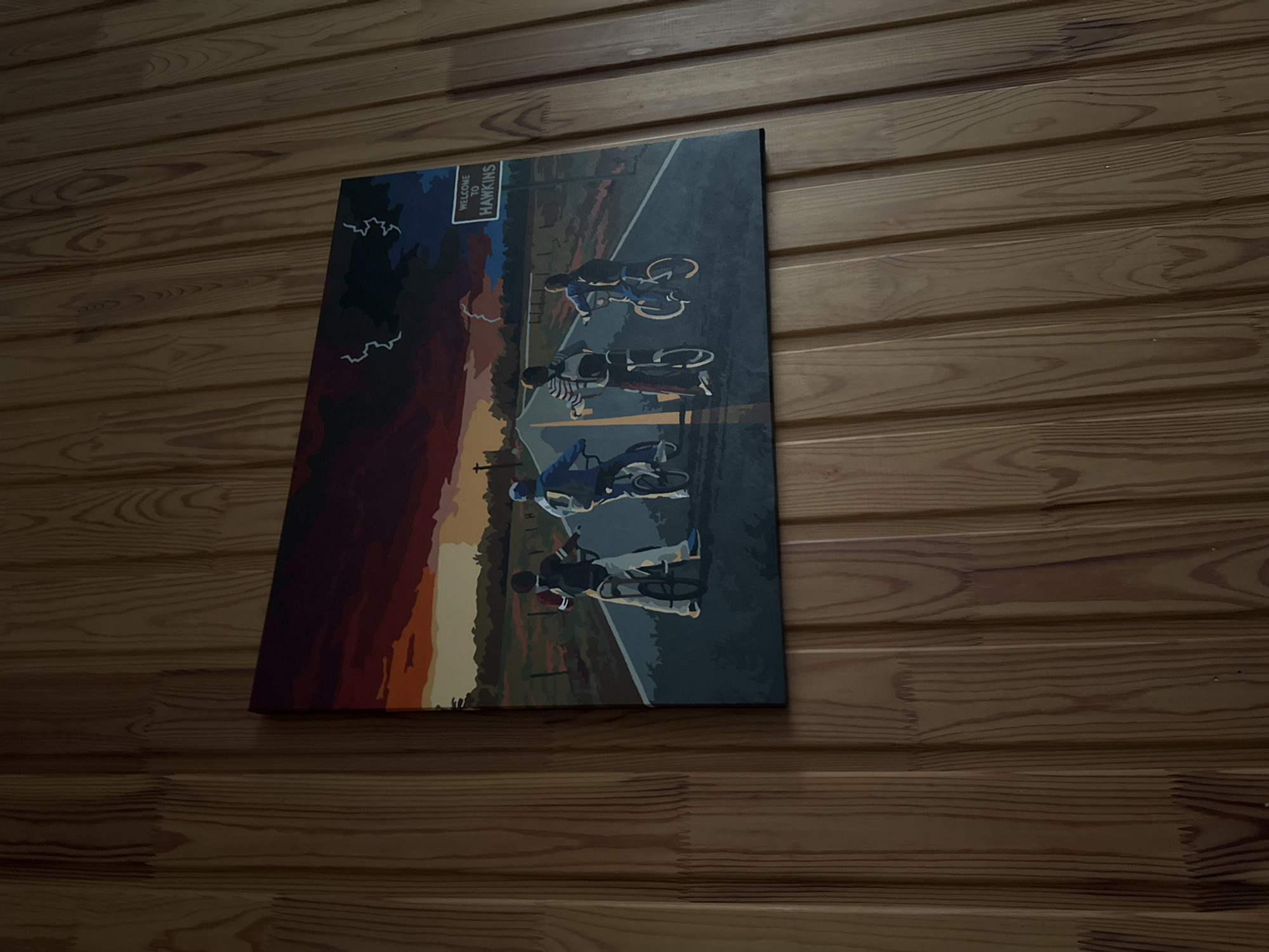 The image displays a rectangular canvas art piece depicting four children on bicycles riding down a road. The artwork has a dark, stormy sky with hints of lightning and a dramatic sunset. A "Welcome to Hawkins" sign is visible on the right side of the road. The canvas is mounted on a wall covered in vertical wooden planks with a visible grain pattern. The scene is static and illustrative, with no discernible action or interaction occurring. The artwork's style is stylized and graphic. The location of this image is not determinable from the visual information provided.