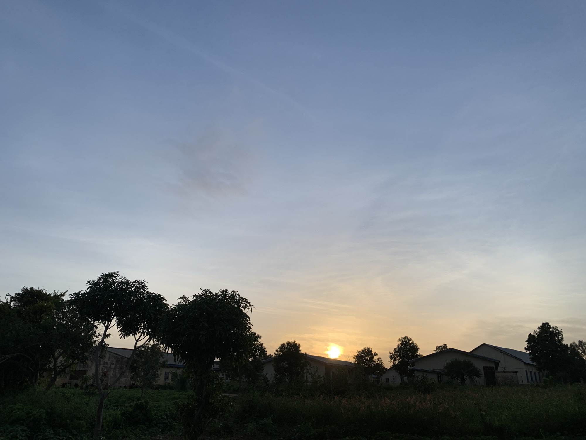 The image depicts a rural scene at dusk, likely in Zawan, Nigeria. The foreground is dominated by a dense collection of trees and foliage silhouetted against the fading light. Behind the vegetation, several low-rise buildings with distinctive pitched roofs are visible, their outlines softened by the low angle of the sun. A single, bright sun, partially obscured by a cloud formation, sits low on the horizon, casting a warm, orange glow that transitions to a pale blue in the upper portion of the sky. The sky itself is a canvas of soft hues, with thin, wispy clouds streaking across it. The overall impression is one of quietude and the end of a day.