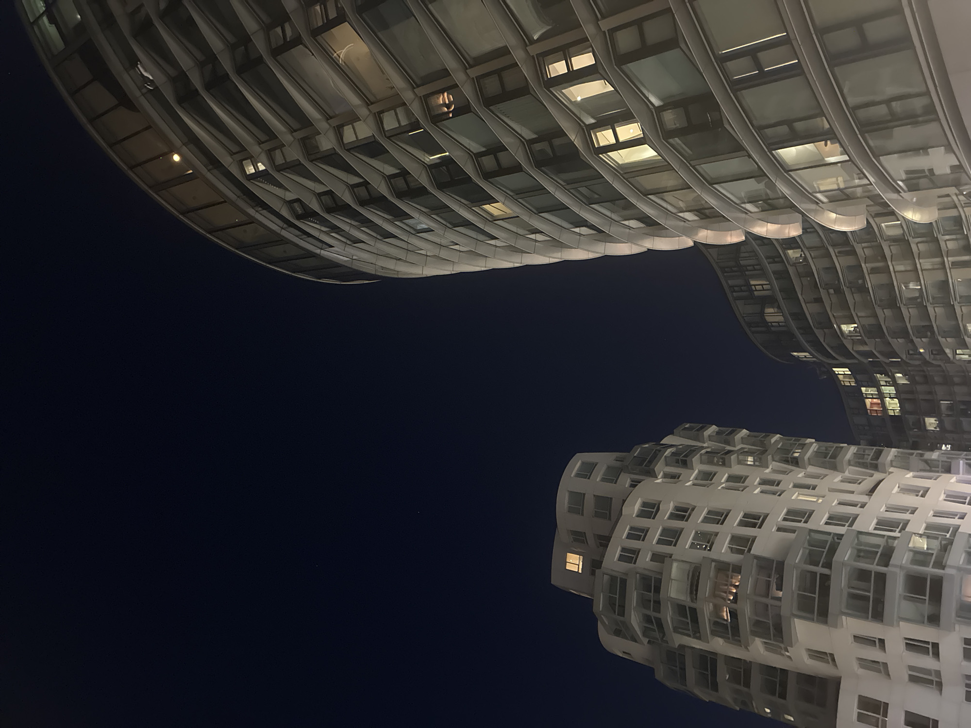 The image shows a night view of two buildings with a distinctive, undulating facade in London, United Kingdom. The buildings are white with numerous windows, some of which are illuminated. The sky is dark. The buildings appear to be located near each other, as their facades seem to curve towards each other. No people are visible in the image.