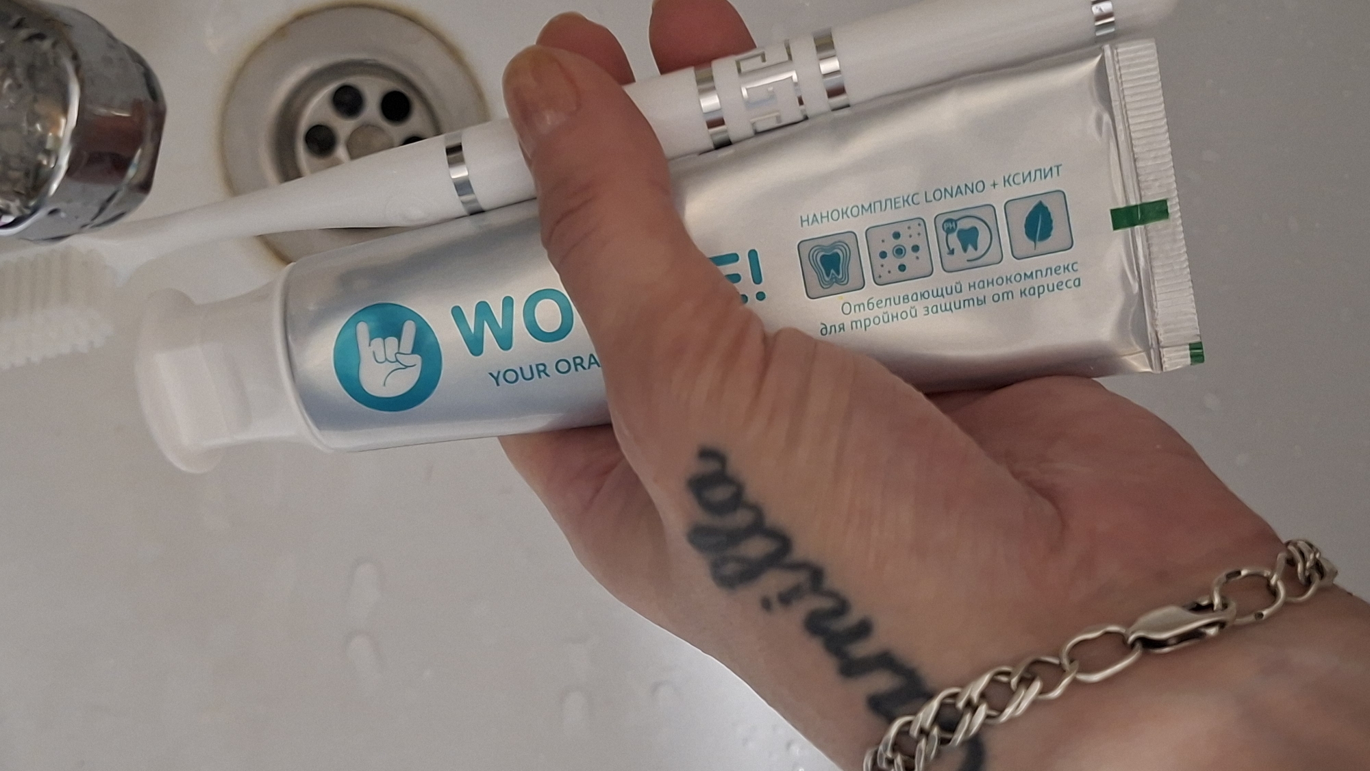 A close-up shot captures a person's hand holding a tube of "WOW! YOUR ORAL CARE" toothpaste and a white toothbrush. The toothpaste tube is silver with blue and white accents, featuring a logo of a hand with a rock-and-roll gesture. Text on the tube is partially visible in Russian, indicating it's for teeth whitening and cavity protection. The toothbrush is white with silver detailing. The hand has a tattoo on the wrist that reads "camilla" in cursive script and is adorned with a silver bracelet. The background appears to be a sink, possibly in a bathroom, with a chrome faucet and drain visible. The scene is well-lit, highlighting the textures of the skin, the toothpaste tube, and the metal fixtures. The setting is indoors and domestic. There is no information in the image to determine the specific location in Забалоцкі сельскі Савет, Belarus.