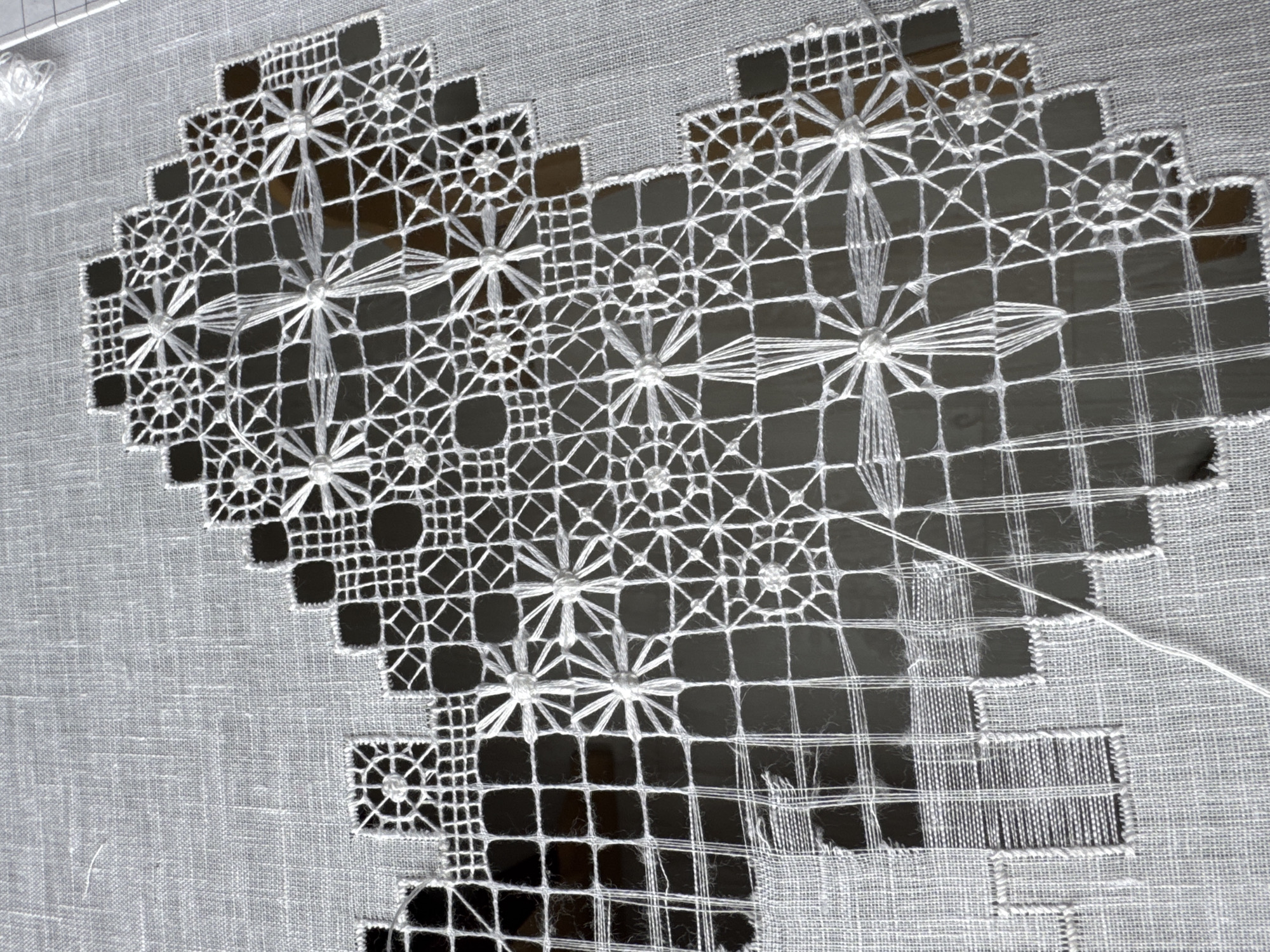 A close-up shot reveals a piece of white embroidery depicting a heart shape. The embroidery features intricate openwork, with geometric patterns, including squares and star-like designs, creating visual interest. Dark voids exist within the pattern, exposing a dark, possibly shadowed, surface beneath. A length of thread extends from the unfinished embroidery. The overall composition suggests delicate craftsmanship and the progress of an ongoing embroidery project. There is no discernible indication of location, specifically Saint Petersburg, Russia, nor are there any people present.
