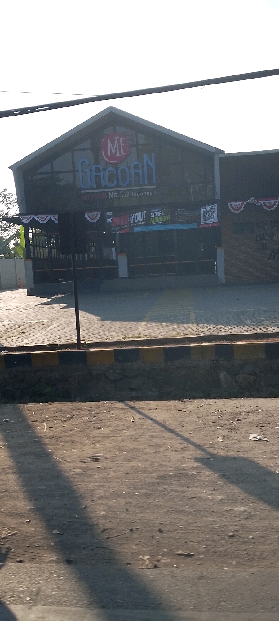The image shows the exterior of a restaurant named "Mie Gacoan" in Bekasi, Indonesia. The restaurant has a distinctive modern architectural design with a dark facade and a prominent sign featuring the restaurant's logo. The sign displays "ME" in a red circular emblem above the word "GACOAN" in large blue letters. Below this, the text "MIE PEDAS No.1 di Indonesia" (Spicy Noodles No. 1 in Indonesia) is visible.
A banner advertising for a "STORE MANAGER" is displayed on the front of the building, with the text "WE NEED YOU! are you ready to grow with us". A QR code is also present on the banner. Decorative red and white pennant banners are hung across the front of the building.
The foreground of the image shows a paved area, possibly a parking lot, with yellow and black striped markings and a low stone wall. The ground is covered in dirt and debris, and a long shadow stretches across it, indicating a sunny day. In the background, trees and a fence are partially visible. The overall impression is a commercial establishment in an urban or semi-urban setting in Bekasi.