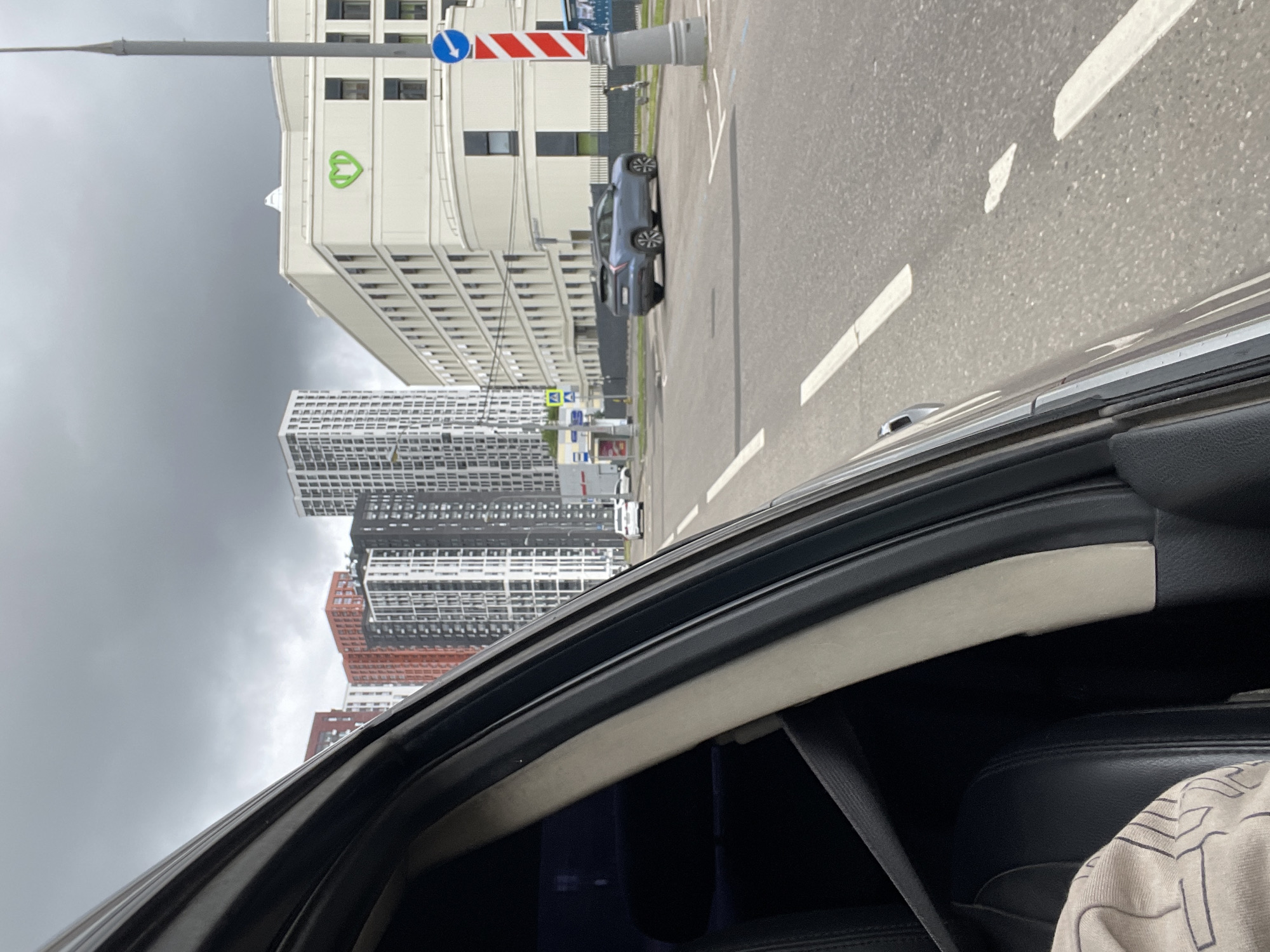 This is a first-person perspective shot from inside a vehicle looking out at a street scene in Moscow, Russia. The view is dominated by the interior of the car on the left, showing a black leather seat and a seatbelt. The exterior view includes a modern apartment complex with tall buildings, some with red and orange facades, and others with white and gray geometric patterns. Across the street is a light-colored building with rounded corners and a prominent green heart logo. A light blue car is parked in front of this building. Further down the street, there are more tall residential buildings. The sky is overcast with dark, heavy clouds. Road markings, including lane dividers and pedestrian crossing symbols, are visible on the asphalt. A traffic pole with red and white stripes and a blue directional arrow sign is also present. The overall atmosphere suggests a cloudy day in a developed urban environment.