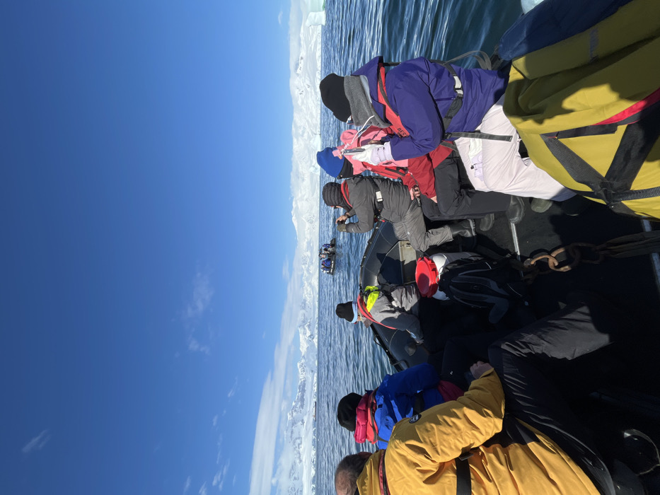 A group of at least seven individuals are onboard a black inflatable boat, navigating calm, dark blue water under a clear, bright blue sky with sparse clouds. The immediate environment features extensive snow-capped mountain ranges and ice formations along the horizon, strongly indicative of a polar marine environment.
The occupants of the foreground boat are dressed in heavy winter expedition gear, including thick jackets (yellow, blue, grey, red, purple), black beanies, and life vests. One individual in a grey jacket is holding a dark camera to their eye, pointing forward. Another individual in a red jacket is holding a white smartphone horizontally, as if taking a photograph, also pointing forward. A third person, in a purple jacket and white pants, is seated next to the smartphone user. Visible equipment in the boat includes a black backpack, a white barrel with a red lid, and a metal chain.
In the mid-distance, another similar black inflatable boat, occupied by several people, is observed on the water. A large, white iceberg is visible towards the far right of the scene. The overall lighting suggests a sunny day.