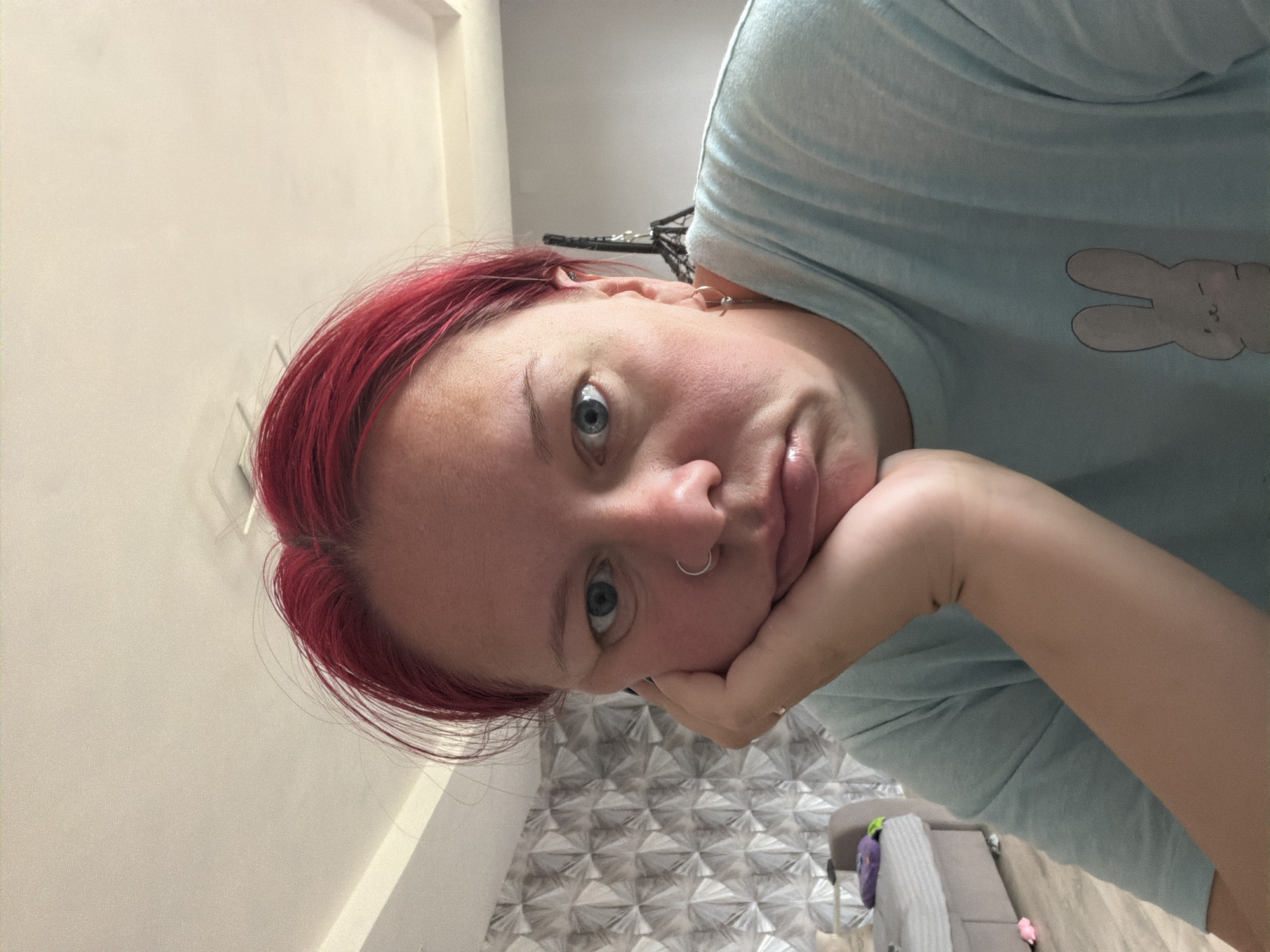 A woman with bright red hair and blue eyes is resting her chin on her hand, looking bored. She is wearing a light blue t-shirt with a drawing of a rabbit on it. She has a nose ring and earrings. The background shows a modern interior with patterned wallpaper and a bed in the corner. The scene appears to be indoors, possibly a bedroom or living room. There is no indication of the city of Chaikovsky, Russia.