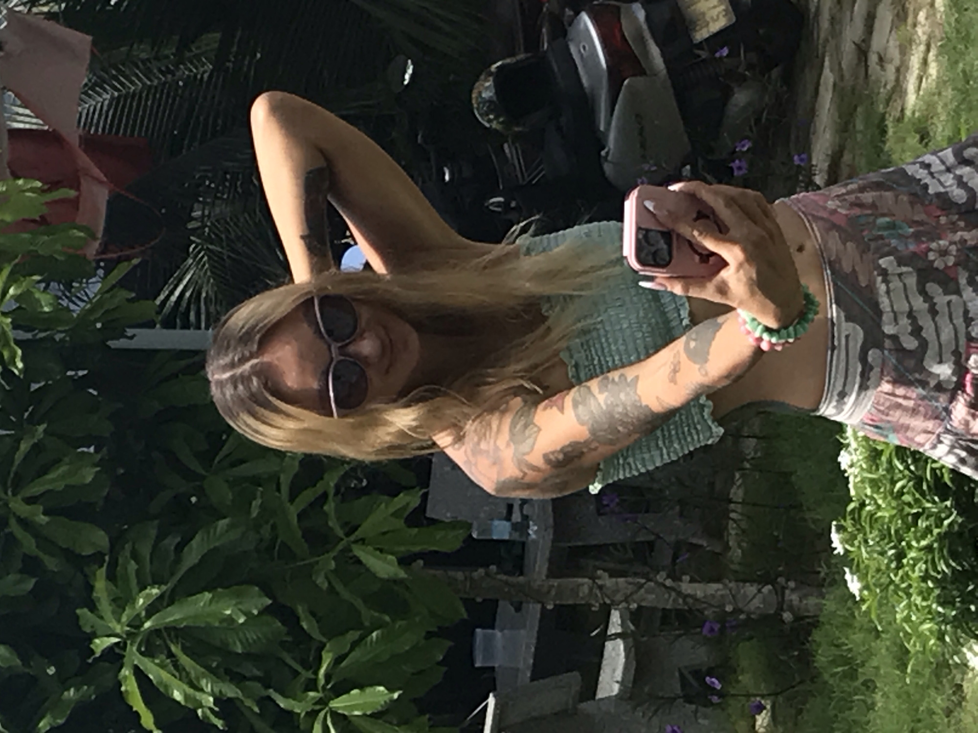 A woman with blonde hair and sunglasses stands in a tropical outdoor setting, consistent with Mũi Né, Vietnam. She is taking a selfie with a pink smartphone held in her left hand, wearing a light green strapless top and a patterned skirt. Tattoos are visible on both arms, and she wears multi-colored beaded bracelets on her left wrist. Her right arm is raised, with her hand positioned near her head. In the background, lush green foliage is prominent, including large-leafed plants and palm fronds. To the left, a light-colored table or bench with containers and a tree trunk wrapped in string lights are visible, with purple flowers on the ground. To the right, a dark-colored scooter with a helmet on its seat is parked, and light-colored laundry hangs from a line in the upper right corner. The ground is covered in green grass.