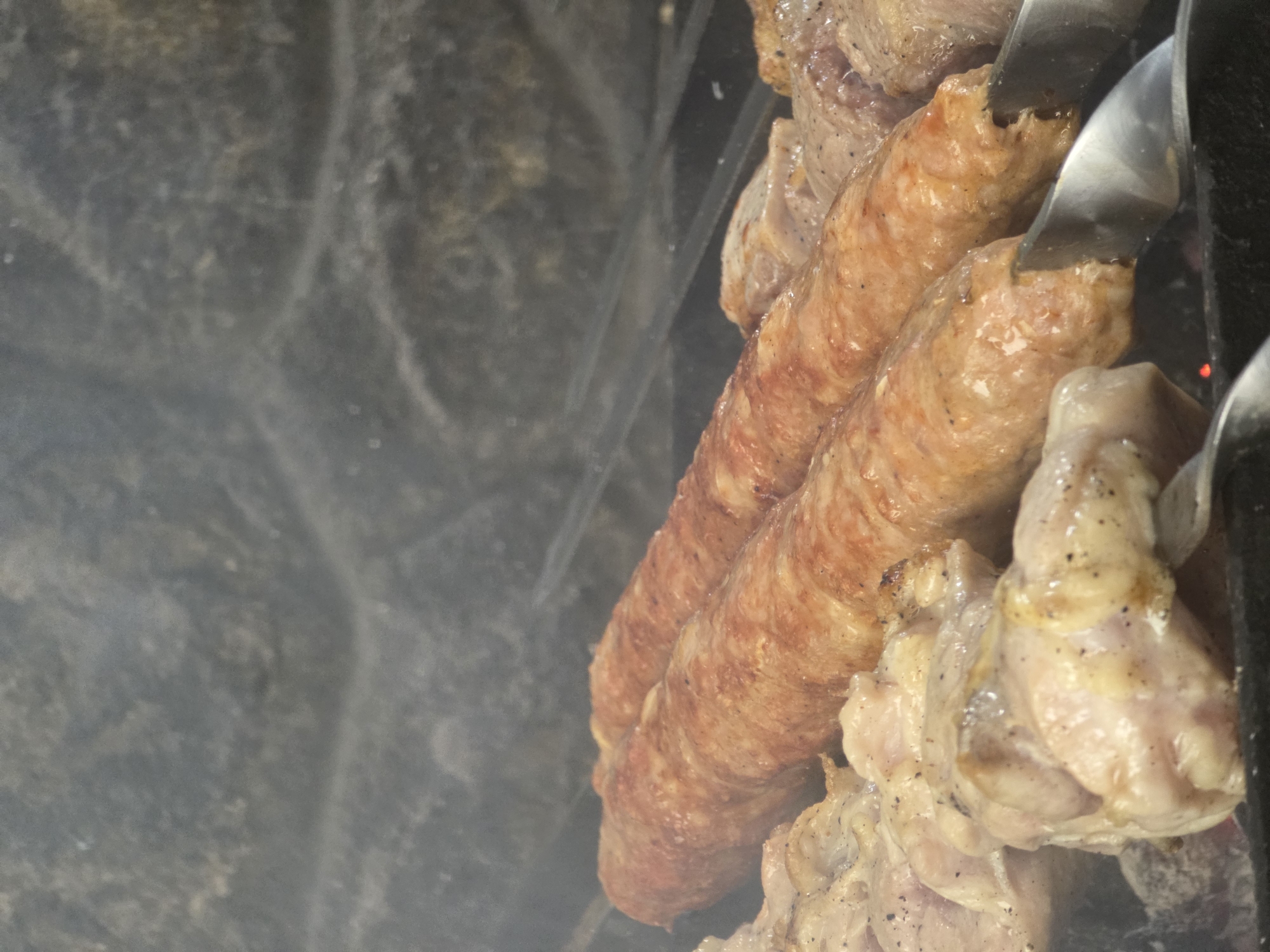 This media file shows a close-up, top-down view of meat grilling on skewers over a dark, textured surface, possibly a grill or fire pit. The main subjects are several pieces of meat, including two large sausages and pieces of what appear to be chicken. The sausages are elongated and have a reddish-brown, slightly wrinkled skin, indicating they are cooked or cooking. They are seasoned with black pepper. The chicken pieces are paler, with visible seasoning and some browning. Metal skewers hold the meat, and part of a metal tool, possibly tongs, is visible, interacting with the sausages. Smoke is present, partially obscuring the background and adding to the impression of a cooking process. The background is a dark, out-of-focus texture, which could be a grill grate or the surrounding environment. There is no visible text in the image. The scene suggests an outdoor cooking event, likely a barbecue or grilling session. The lighting is not indicative of a specific time of day, and there are no weather cues. Given the location context of Pidhorodne, Ukraine, this could be a common outdoor meal preparation. The overall impression is of food being cooked, with a focus on the meat and the grilling process.