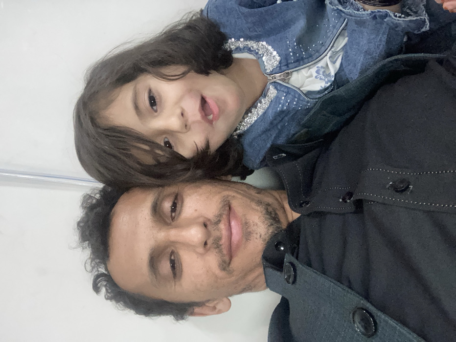An adult male and a young female child are depicted in a close-up indoor portrait in Quetta City Tehsil, Pakistan. The man, positioned on the left, has dark, curly hair and facial stubble, wearing a dark button-up shirt or jacket over a dark undershirt, exhibiting a slight smile. The child, positioned to the right of the man and slightly forward, has dark, shoulder-length hair with bangs and an open-mouthed expression. She is wearing a light blue denim jacket embellished with silver sequins, over a white top with a blue floral pattern. A dark bracelet or bangles are visible on her left wrist. Both individuals are looking towards the camera. The background is uniformly light-colored, with a thin vertical line or object visible in the upper right.