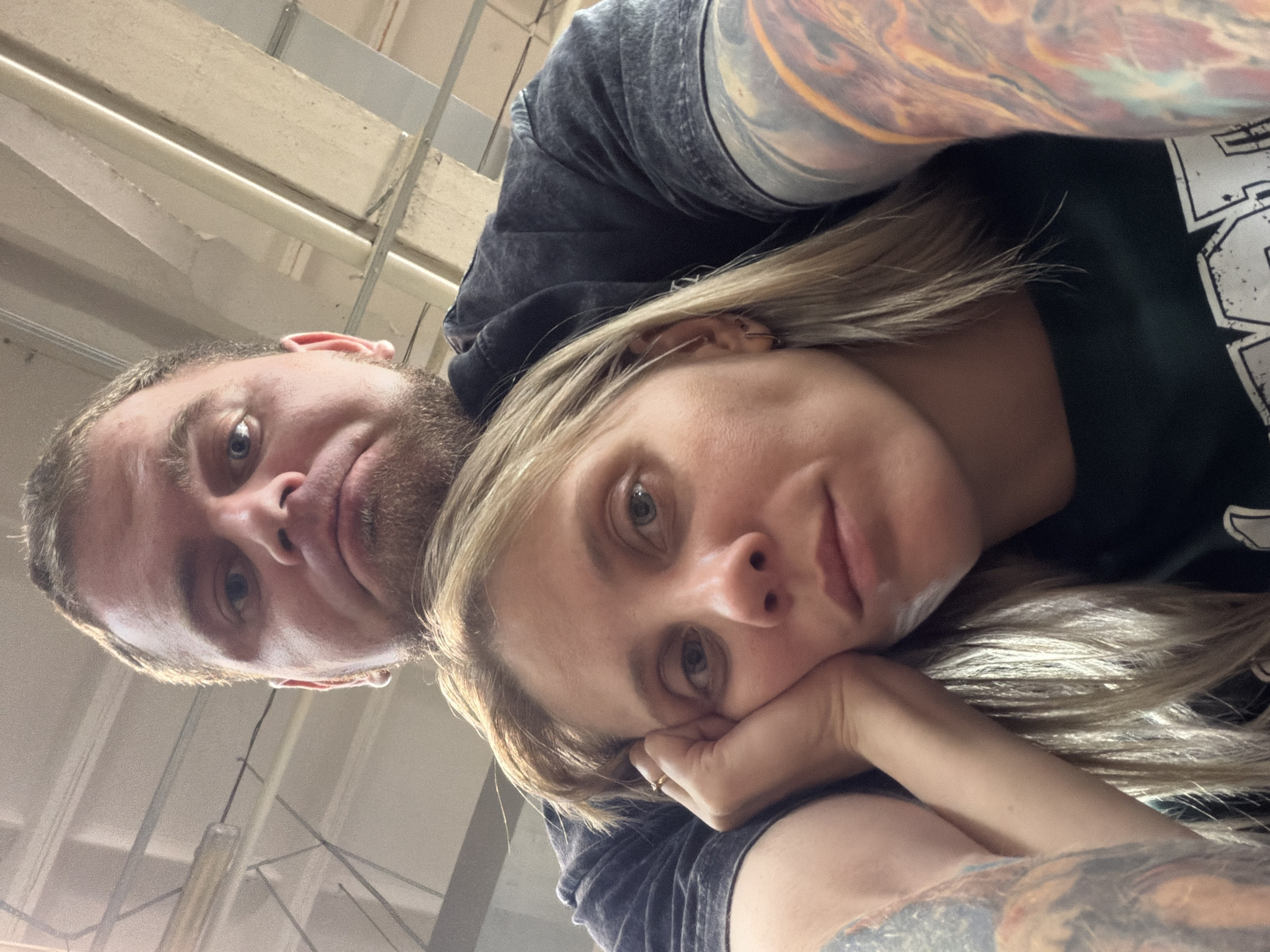The image is a close-up selfie of a man and a woman. The man, positioned behind the woman, is looking down with a slightly downturned mouth. He has short brown hair and a beard. The woman, in the foreground, is resting her head on her hand and looking towards the camera with a tired expression. She has blonde hair and visible earrings. Both individuals are wearing dark t-shirts. The background appears to be an indoor space with a ceiling structure that includes beams and fluorescent lighting, possibly a warehouse or industrial building. The lighting suggests an overcast day or artificial illumination. The location is identified as Chaikovsky, Russia.