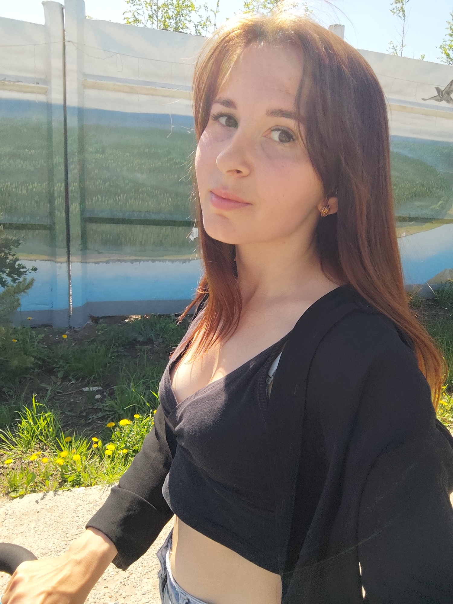 The image is a medium close-up shot of a fair-skinned woman with reddish-brown hair. She is wearing a black crop top and a black cardigan, paired with denim jeans. The background includes a concrete wall painted with a mural depicting a grassy landscape with a blue sky. Some dandelions are visible in the foreground next to a concrete path. The location is Chaikovsky, Russia.
