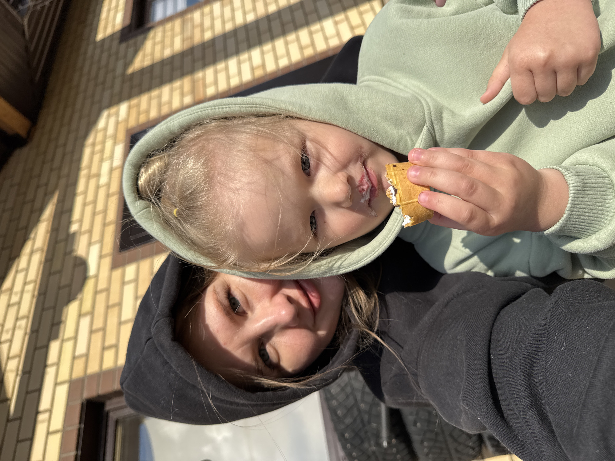 The image captures a close-up of a young girl and an adult, possibly her mother, in what appears to be Gornyi, Dvurechenskoe S/P, Russia. The girl is in the foreground wearing a light green hooded sweatshirt and holding a waffle ice cream dessert. Some of the ice cream is on her face. The adult behind her is wearing a black hooded sweatshirt. Brick building is in the background.