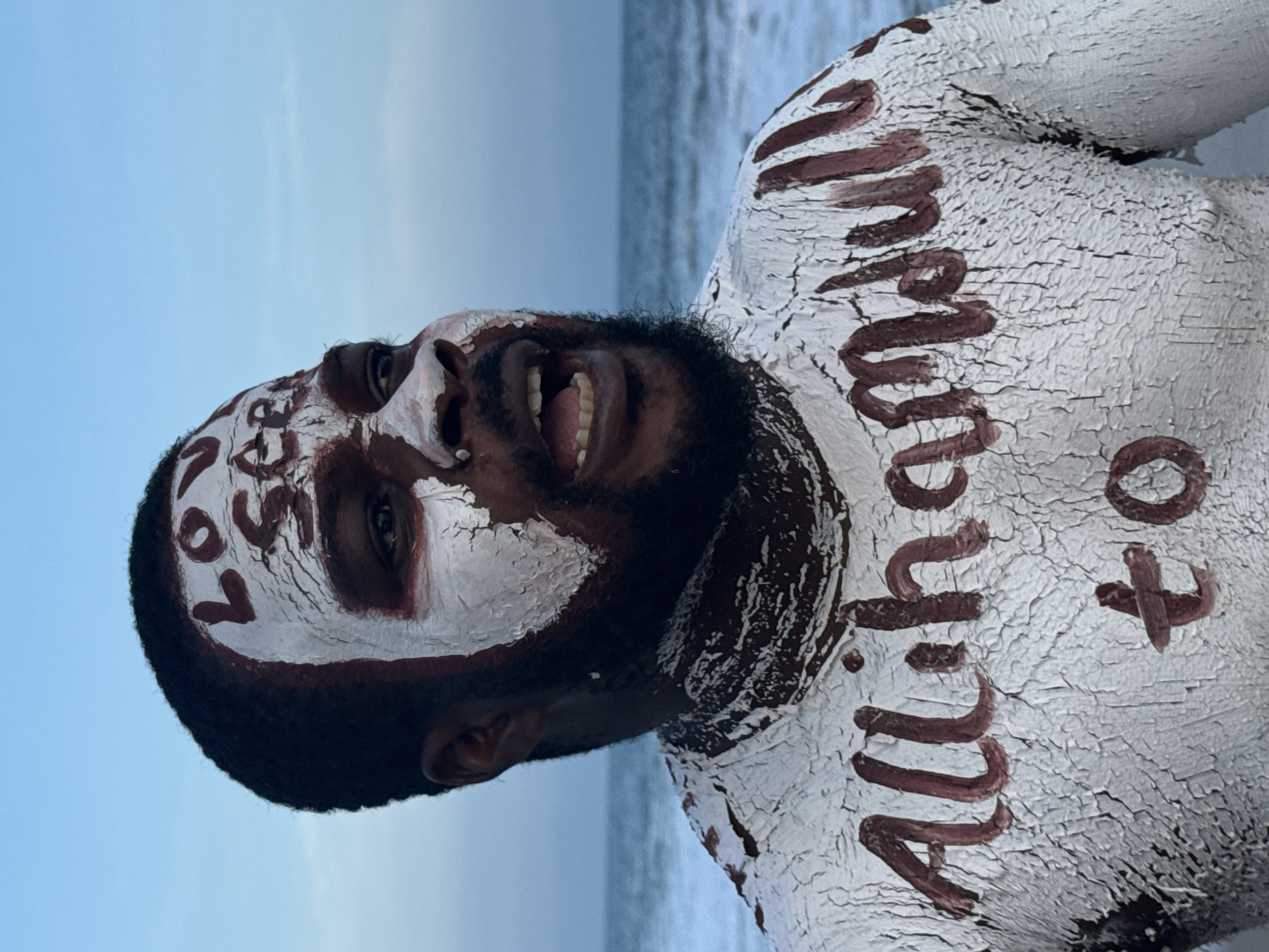 A person with dark skin and a black beard is standing near the beach in Mopo Onijebu, Nigeria. Their face and torso are covered in cracked, white paint with brown writing. On the face, the word "LOVE" is written on the forehead and "LOSS" on the cheek. On the torso, partial words appear to read "to All..."
The person is looking upwards with their mouth open. The backdrop includes the beach and the sky.