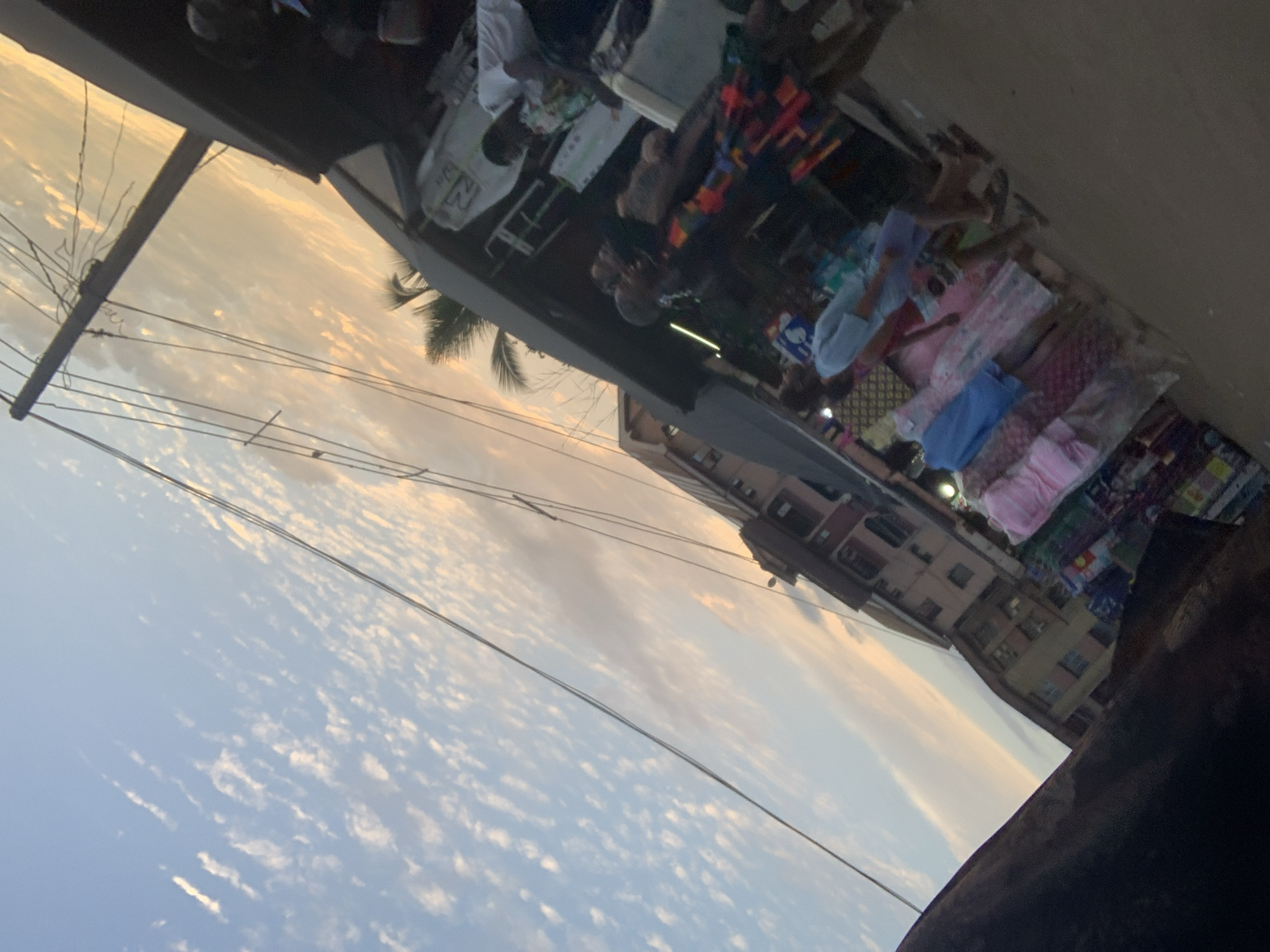 The image captures a street market scene in Obosi, Nigeria, likely during the late afternoon or early evening given the warm hues of the sky. The perspective is tilted, giving a dynamic feel to the composition. In the foreground, a dark, weathered surface, possibly corrugated metal or tarpaulin, occupies the bottom left.
Dominating the upper portion of the image is a vast sky filled with a delicate pattern of cirrus clouds against a pale blue background. The light suggests either sunrise or sunset, casting a warm, orange glow on the clouds and the horizon. Several power lines crisscross the sky, supported by a utility pole visible on the right side.
Below the sky, the scene transitions to the market. Buildings line the street, their architecture suggesting a tropical or sub-tropical urban environment. The structures appear multi-storied with visible windows and balconies. A palm frond is visible peeking from behind one of the buildings.
The market itself is bustling with activity. Stalls display a variety of goods, primarily clothing. Garments, including dresses and what appear to be shirts, are arranged in a colorful display, some neatly folded and others hanging. Several people are present, engaged in shopping or attending the stalls. Their clothing styles and skin tones are consistent with the region. One individual, seen in the middle ground, wears a brightly colored, patterned dress. Another person, partially obscured, is wearing a white t-shirt. The ground appears to be a dirt or paved surface typical of a market environment. The overall atmosphere is one of lively commerce and daily life in Obosi.