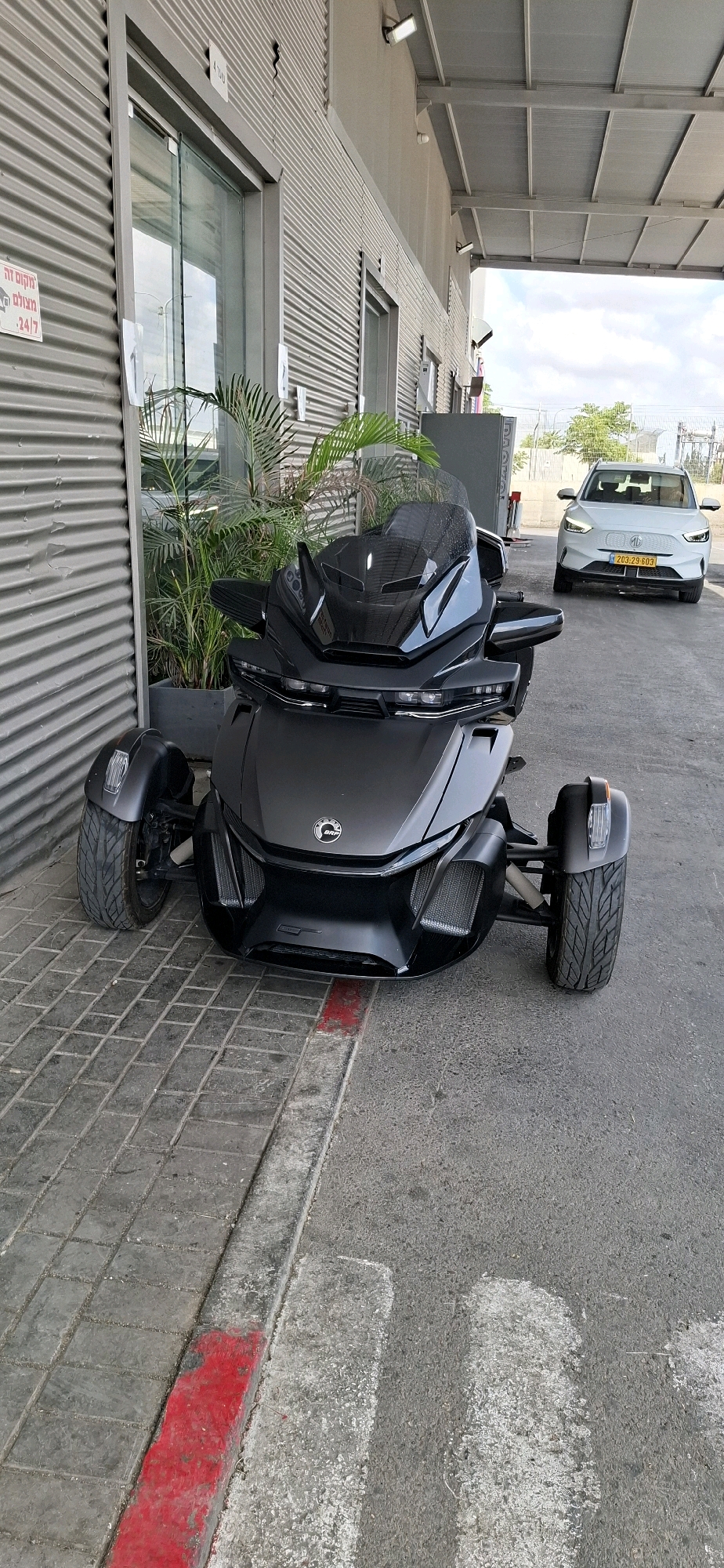 A black Can-Am three-wheeled motorcycle is parked on a cobblestone walkway next to a corrugated metal building. A white electric vehicle with license plate "203-29-603" is visible parked in the background. The building has large glass doors and windows. In front of the motorcycle is a large potted plant with palm-like fronds. The walkway has a red painted curb. The scene appears to be in an industrial or commercial area, likely in Kiryat Gat, Israel, given the Hebrew text on a sign visible on the building. The sky is cloudy.