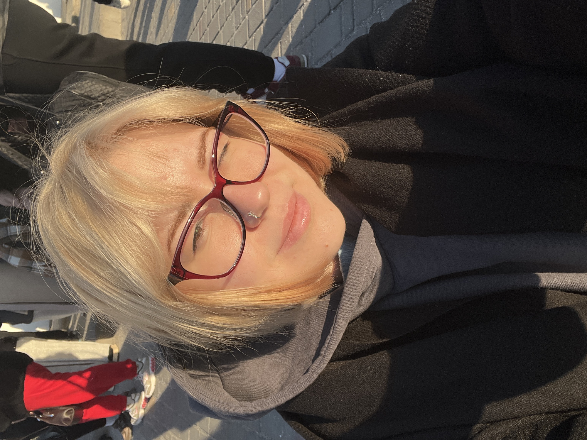 A pale-skinned person with blonde hair and red-framed glasses is the primary subject of this image, possibly a selfie. They are wearing a dark coat over a grey hooded sweatshirt. A nose ring is visible. The background reveals a paved surface, with other individuals in the periphery, some wearing red pants and sneakers. The location appears to be an outdoor public space, possibly in a city.