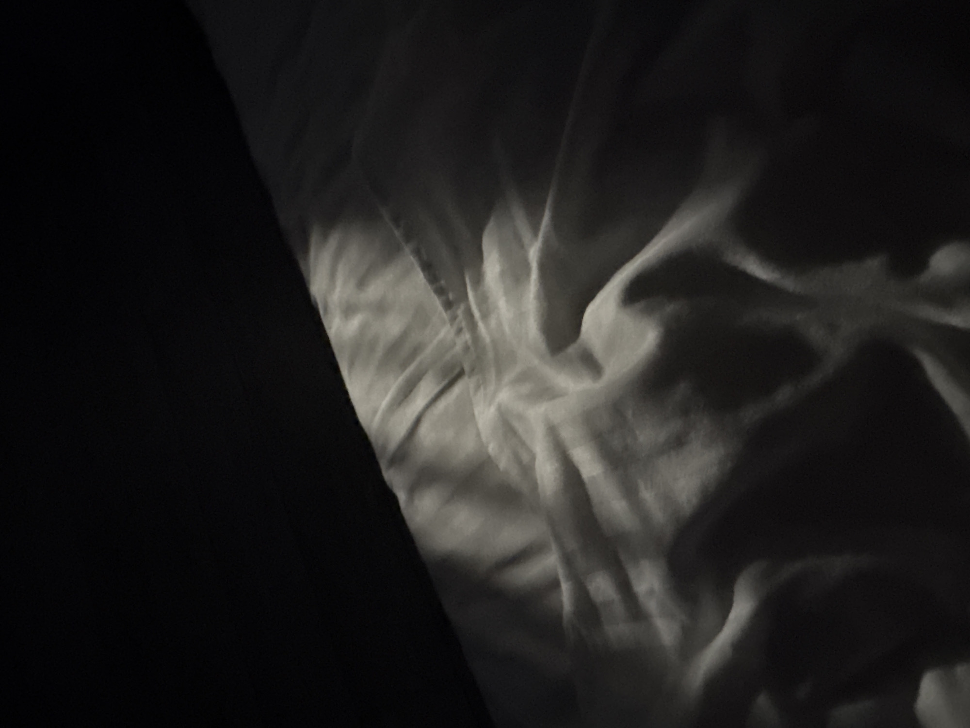 A close-up, high-angle shot captures a crumpled white sheet of fabric, likely bedding, bathed in dramatic chiaroscuro lighting. A dark, shadowed area dominates the top left quadrant of the image, with a sharp diagonal line of light illuminating the texture of the fabric. The fabric itself is creased and folded, creating deep shadows and bright highlights that emphasize its soft, pliable nature. The overall impression is one of stillness and quietude, with the interplay of light and shadow suggesting a sense of depth and volume within the folds of the material. The image provides no contextual information about its location in Doma 1096 Km, Russia, nor does it depict any people, objects, or actions.