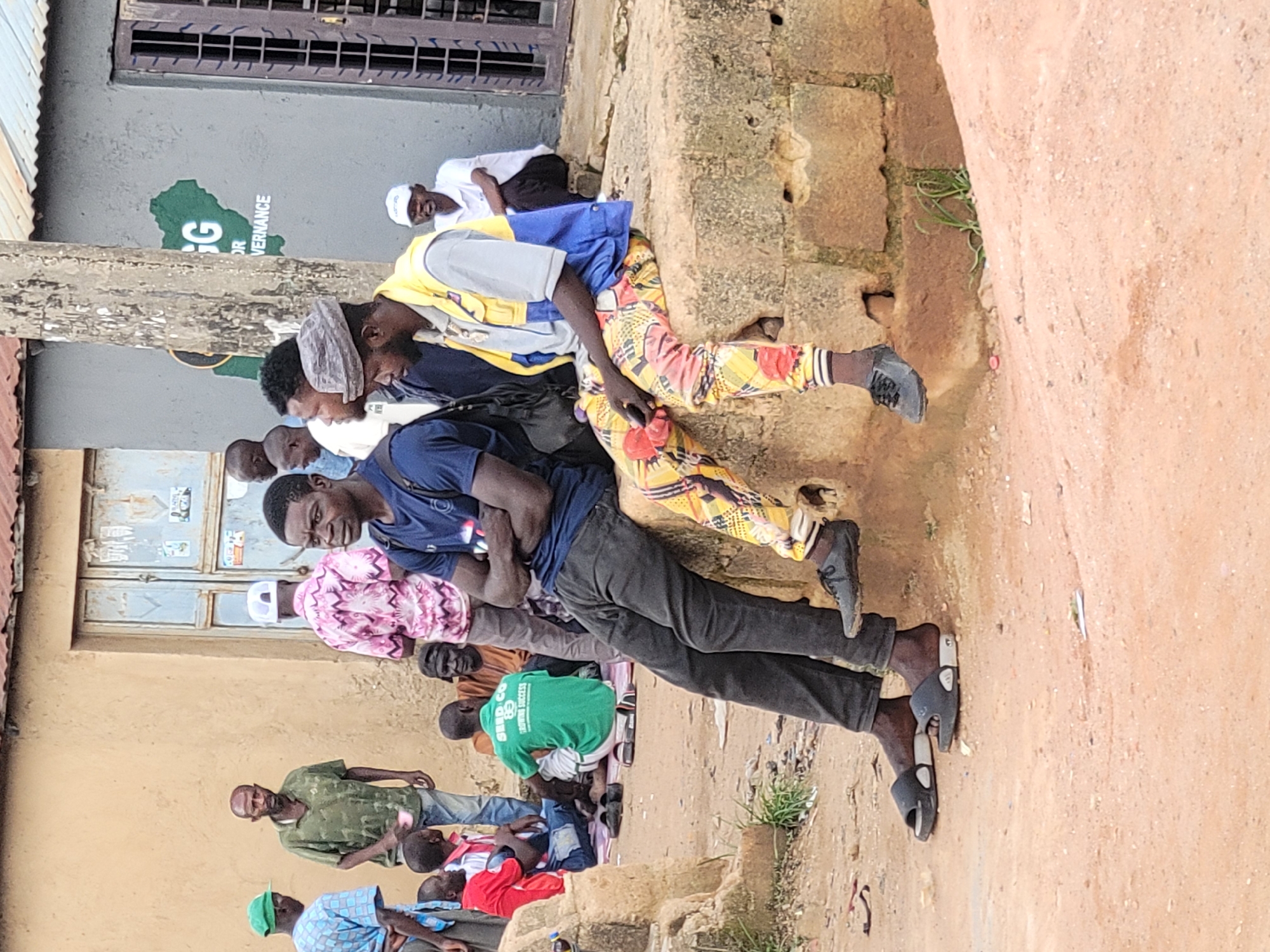 A group of men and boys are gathered on a dusty street outside a building in Jos, Nigeria. Two men are sitting on a low concrete wall in the foreground. The man on the left is wearing a dark blue t-shirt and black pants, with black slide sandals. The man on the right is wearing a yellow and blue vest over a grey shirt, with brightly patterned pants and dark sneakers. Other individuals are standing and sitting in the background. One man in a green shirt is holding a small cup. A building with a faded sign featuring the Nigerian map and the word "GOVERNANCE" is visible behind them. The environment appears to be an urban or semi-urban area with a simple, utilitarian aesthetic.