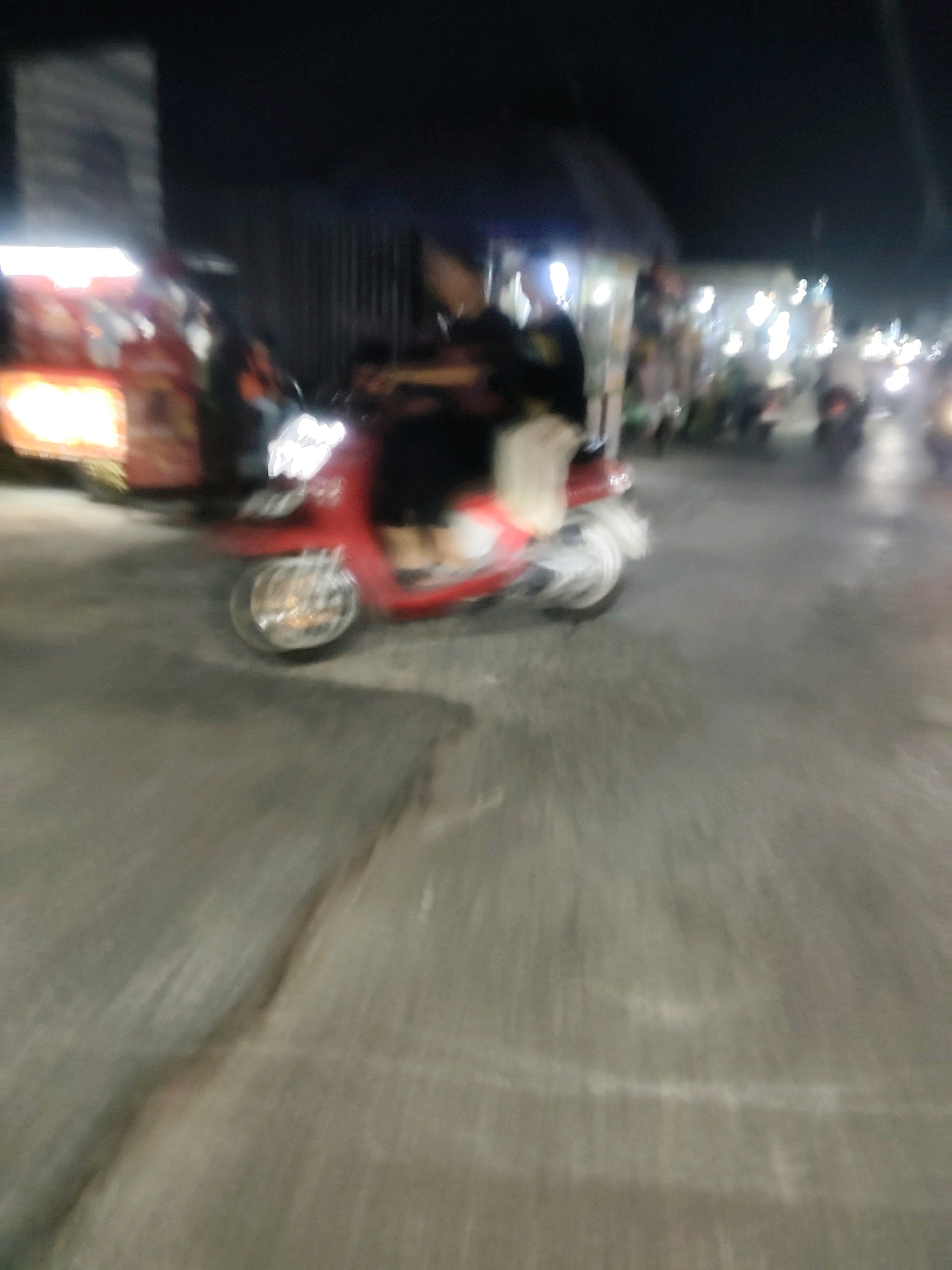 In Mekarmukti, Indonesia, a nighttime scene unfolds with blurry motion and indistinct figures. A red scooter, with a rider partially visible, is the central subject, its headlight casting a bright, diffused glow. The scooter is in motion on a paved road, evident from the blurred asphalt and faint road markings. In the background, the lights of market stalls or shops create streaks of illumination against the dark sky. The overall impression is one of a busy, albeit blurry, street scene at night in an Indonesian urban or semi-urban environment.