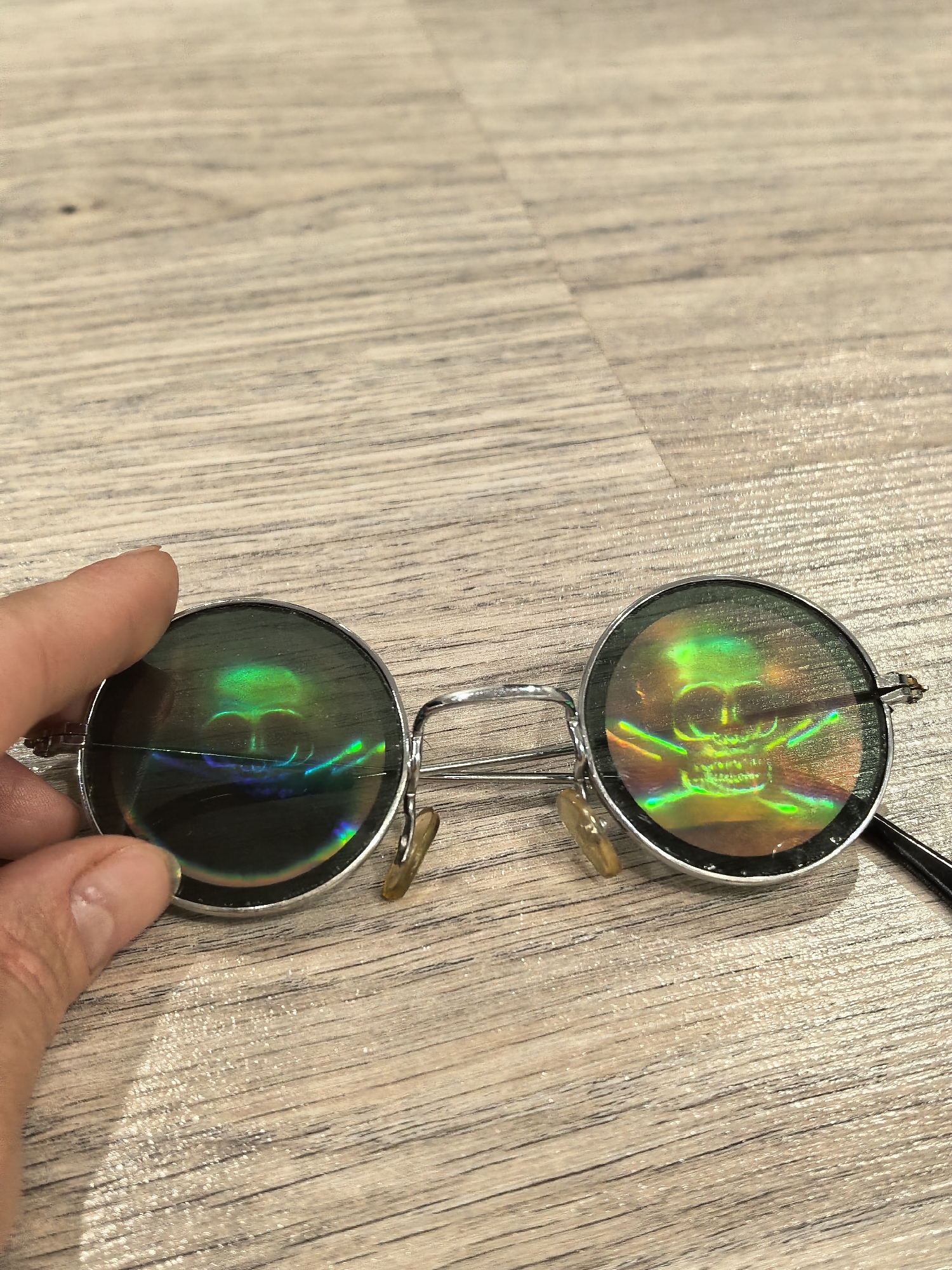 This is a close-up, overhead shot of a pair of round, silver-rimmed sunglasses with holographic lenses resting on a light-colored wooden surface. The lenses display a skull and crossbones motif in vibrant green and rainbow hues. A human hand, with fingernails painted a neutral color, is visible on the left, holding one of the lenses. The background is a wooden surface with a visible grain pattern, suggesting a floor or tabletop. The image does not contain any identifiable landmarks or elements that would suggest a specific location within Sterlitamak, Russia.