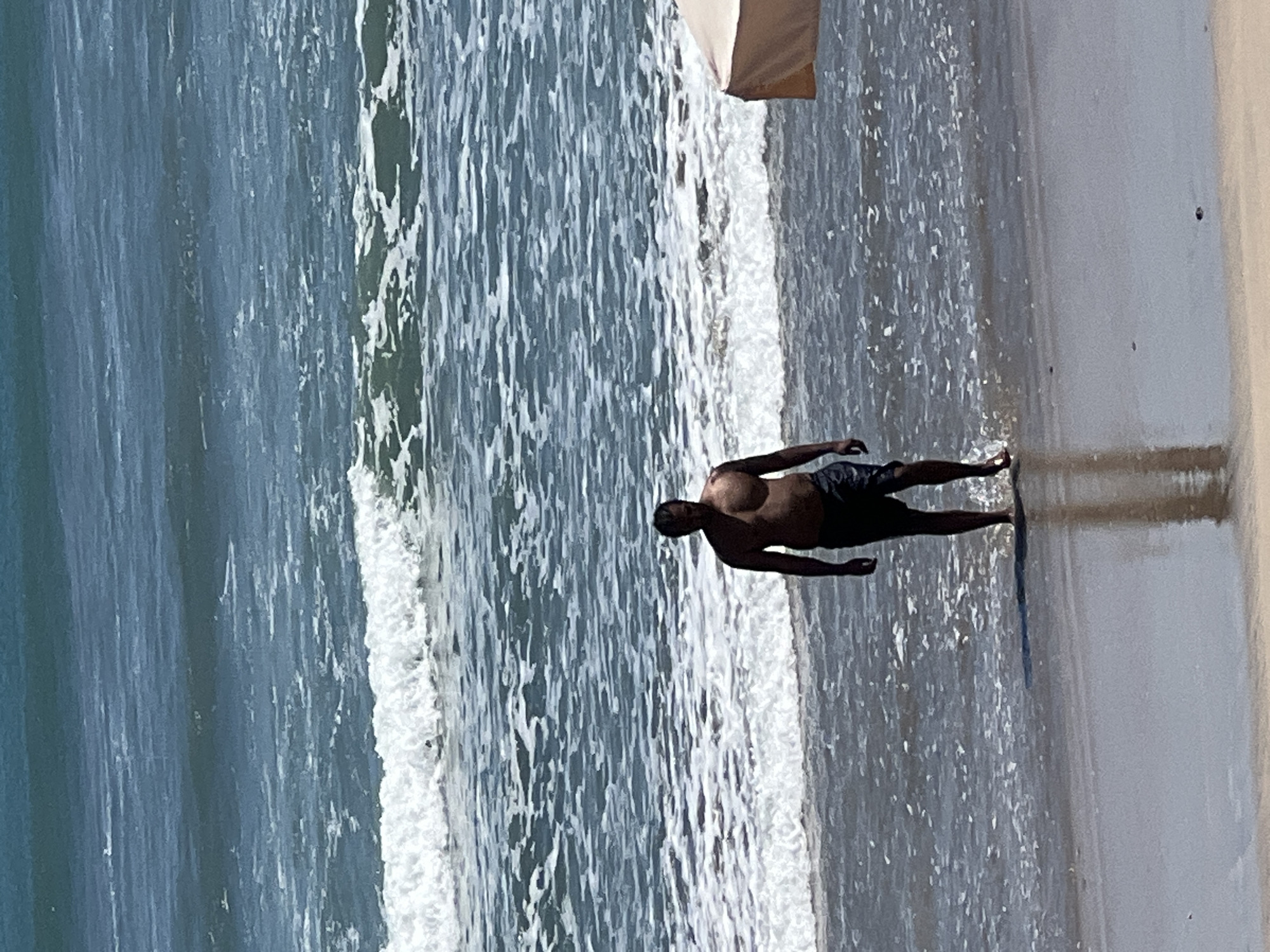 A shirtless man wearing dark swim shorts stands in shallow water at the edge of the ocean. He is facing forward, looking towards the camera, with his arms at his sides. The ocean behind him has moderate waves breaking and forming white foam. The sand in the foreground is wet, reflecting the man and the sky. To the right, a portion of a tan umbrella is visible. The scene suggests a sunny day at the beach in Kuta, Indonesia.