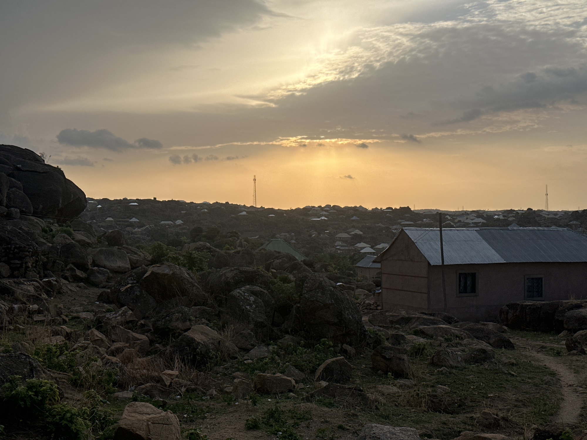 The image shows a landscape in Jos, Nigeria, during what appears to be sunset. The scene is dominated by rocky terrain interspersed with vegetation. A modest, single-story building with a corrugated metal roof stands to the right of the frame. In the background, a densely packed residential area is visible, with similar low-rise buildings stretching toward the horizon. A communication tower is discernible in the distance. The sky is overcast with the sun obscured behind the clouds, casting a diffused, golden light across the scene.