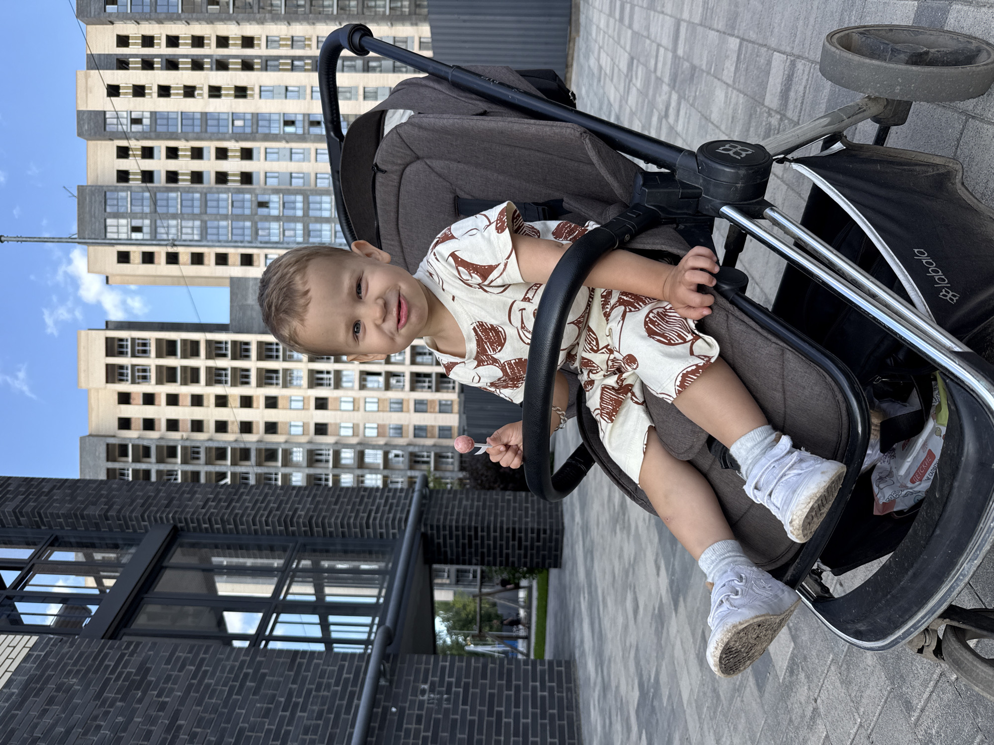 A young boy sits in a stroller on a paved pathway in front of a modern apartment building. The boy is wearing a light-colored outfit with a brown print and white socks and sneakers. He is holding a pink lollipop. The stroller is dark gray with a black handle. In the background, there are tall apartment buildings with many windows. The sky is blue with some clouds. The setting appears to be an urban residential area.