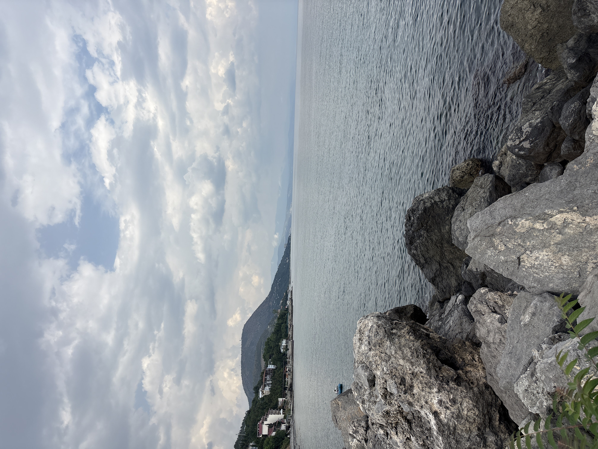 The image depicts a coastal scene in Bondarenkovo, Ukraine. In the foreground, a rocky shoreline is visible, with large grey stones dominating the lower portion of the frame. The sea stretches out to the horizon, its surface rippled by a gentle breeze. The water is a muted grey-blue color, reflecting the overcast sky. In the mid-ground, a small blue boat with a person onboard is visible on the water, moving away from the shore. Along the coastline, a town or village is nestled on the slopes of a hill. Buildings with white and red roofs are interspersed with lush green trees. The hill rises behind the settlement, its peak partially obscured by clouds. The sky is filled with a dense layer of grey clouds, with occasional patches of brighter blue peeking through. The overall atmosphere is calm and serene.