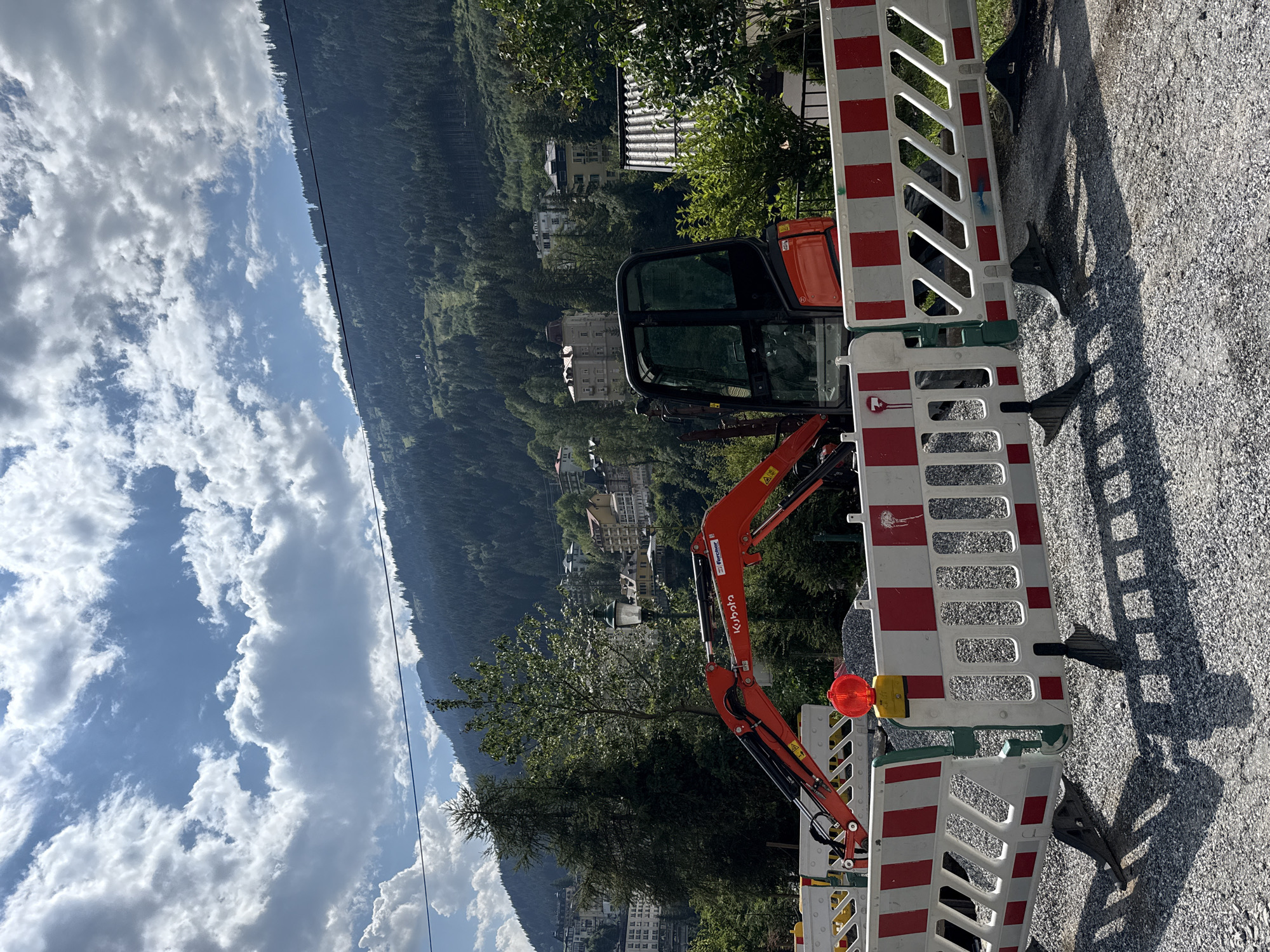 An orange excavator with "Kubota" and "Fe" logos is parked on a gravel surface, partially obscured by a red and white construction barrier. A red flashing light is mounted on the barrier. In the background, a wooded mountain slopes upwards, dotted with buildings. The sky is blue with scattered white clouds. The scene is set in a town, likely Bad Gastein, Austria, as indicated by the mountainous terrain and architecture. The image appears to be taken during daytime, under sunny conditions. There are no people visible in the image.