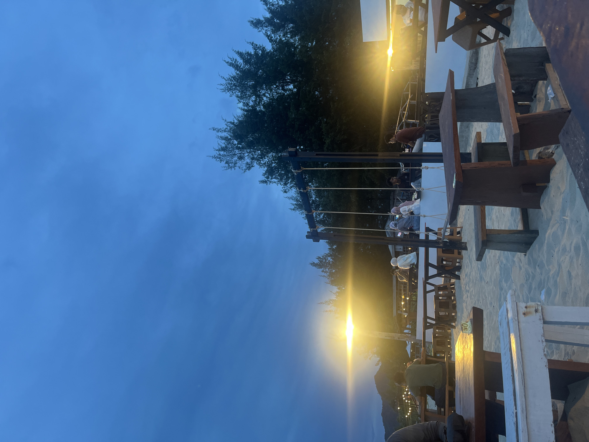 A wide-angle, low-angle shot captures an outdoor seating area on a sandy beach at dusk. The sky is a deep blue with some dark clouds, and a bright, horizontal streak of light, possibly from a setting sun or artificial lighting, illuminates the scene. Several wooden tables and benches are scattered across the sand, some occupied by people. In the background, trees and structures suggestive of a beachfront establishment are visible, along with distant hills. A swing set structure is also present. The overall atmosphere is relaxed and communal.