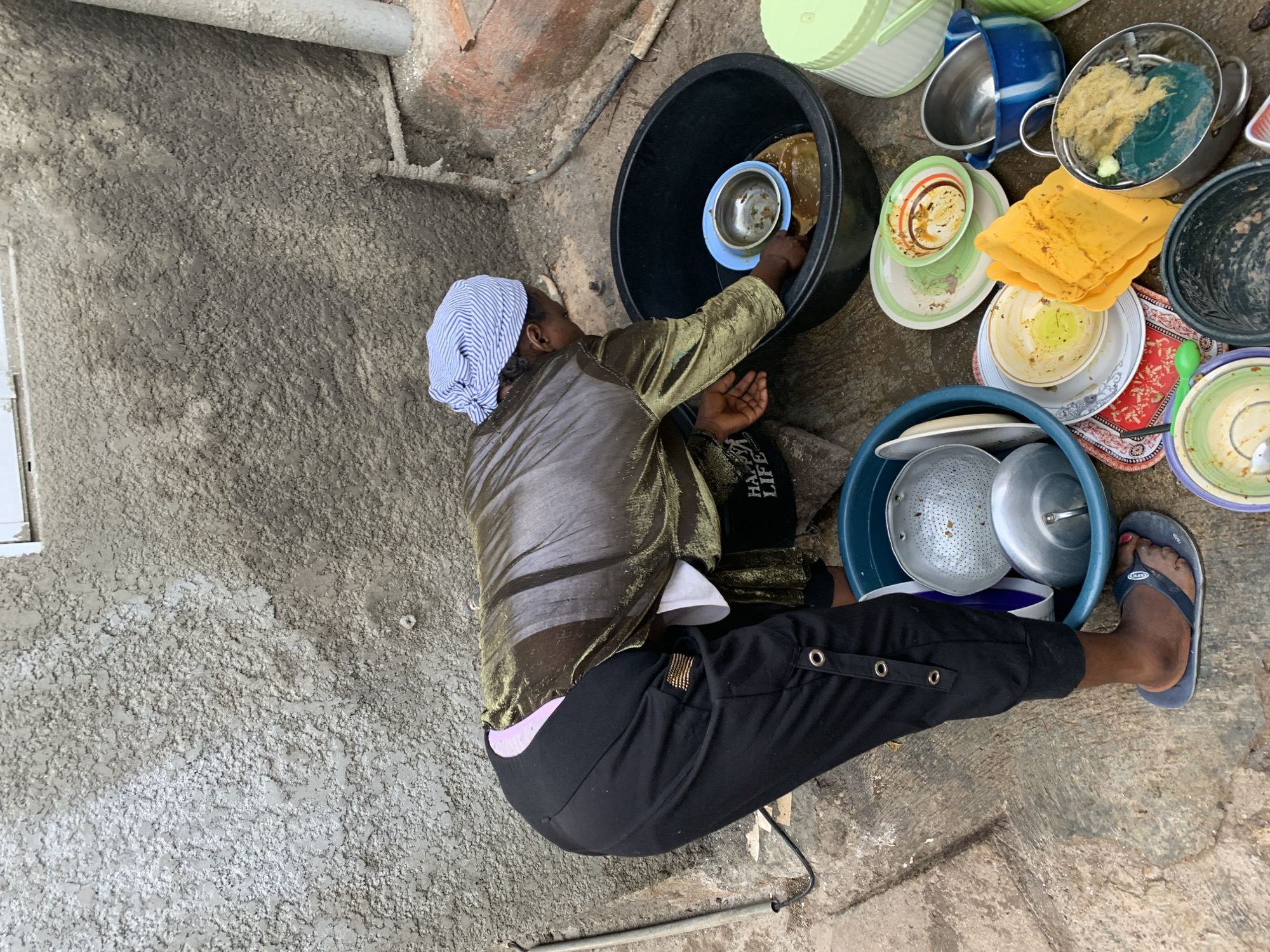 An individual is seated on a concrete surface in an outdoor setting, seemingly engaged in washing dishes. They are wearing a blue and white head covering, a metallic gold-colored shirt, black pants, and a blue flip-flop on their right foot. The individual is seated next to a large black basin containing dishes. Various dirty dishes are stacked around the black basin, including plates, bowls, and pots. The surrounding environment appears to be a courtyard with a textured concrete wall in the background. The location is Jos, Nigeria.