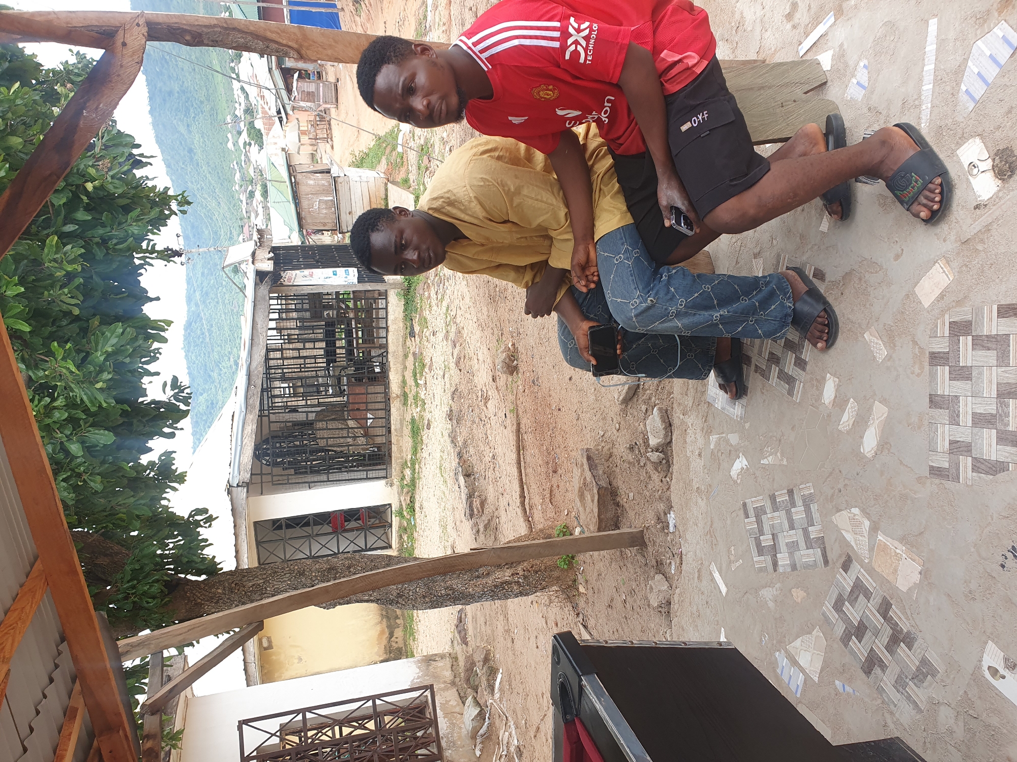 Two men are seated outdoors under a wooden roof. The man on the left wears a mustard-colored traditional garment and blue jeans. He is holding a smartphone. The man on the right wears a red Manchester United jersey and black shorts. He is also holding a smartphone. They are seated on a bench. The ground is made of sand and scattered tiles. Behind them, there are buildings and a tree. The background shows a hillside with scattered buildings. This scene is located in Lokoja, Nigeria.