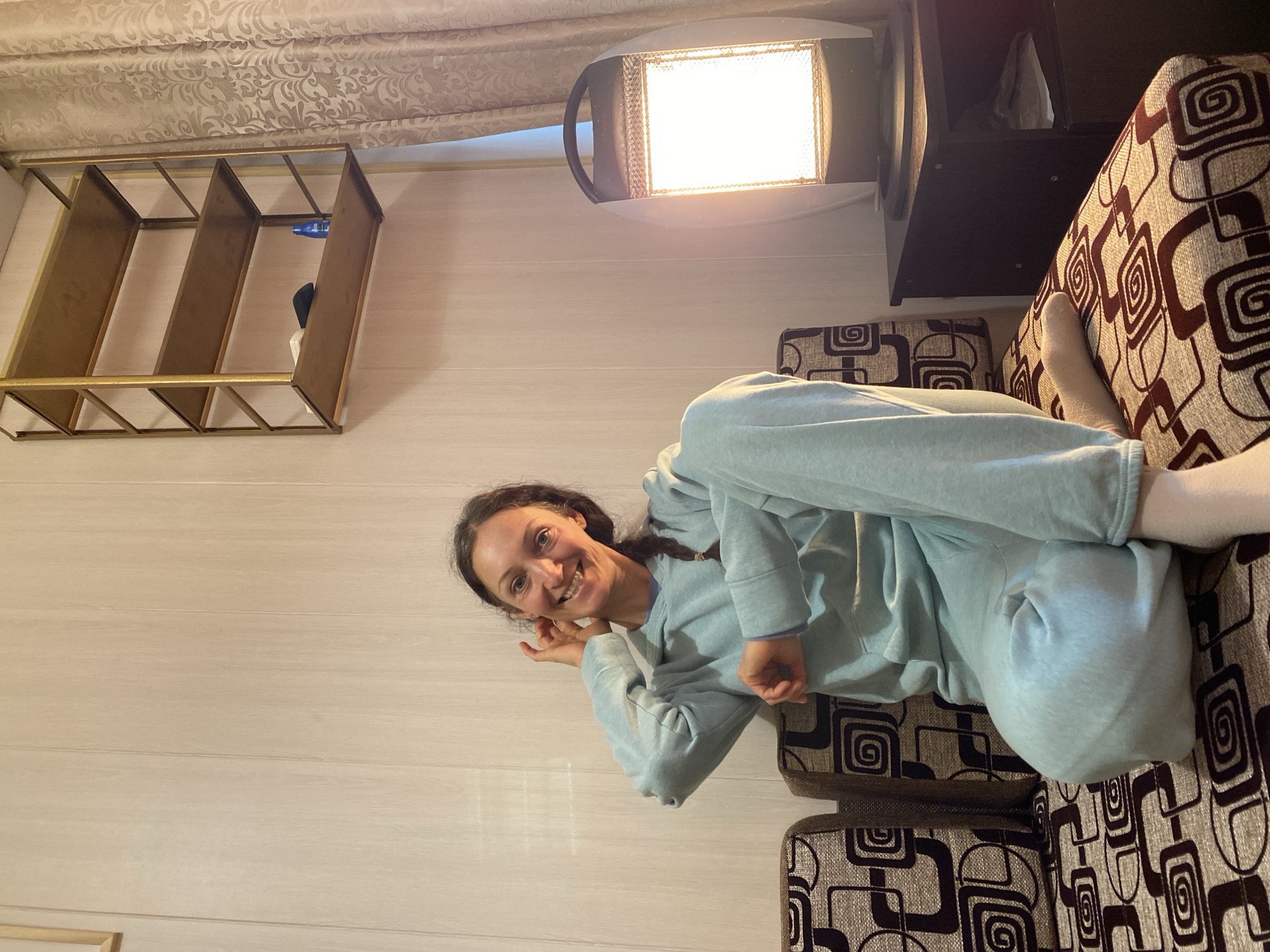 An indoor scene captured in Gelendzhik, Russia, features a woman relaxing on a patterned sofa. She is lying on her back, head turned towards the camera, and smiling broadly, conveying a sense of happiness and ease. Her dark hair appears to be in a braid, and she is dressed in a light blue tracksuit, comprising a hooded top and matching trousers. One of her feet, covered in a white sock, is visible, resting on the sofa's arm or backrest.
The sofa is upholstered with a distinctive brown and beige fabric, featuring repeating geometric and spiral patterns. The walls in the room are clad in light-colored horizontal wooden panels, contributing to a cozy ambiance.
Above the woman's head, a bright, rectangular heater or lamp is mounted on the wall. Its glowing element emits a warm, strong artificial light, suggesting the photo might have been taken during the evening or night. To the left of the light source, a golden-toned open-shelved unit, resembling a decorative shelf or wine rack, is also fixed to the wall. A blue item and a dark object are visible on one of its compartments. In the upper background, light-colored curtains with a subtle floral or damask pattern are draped. No text is visible in the image.