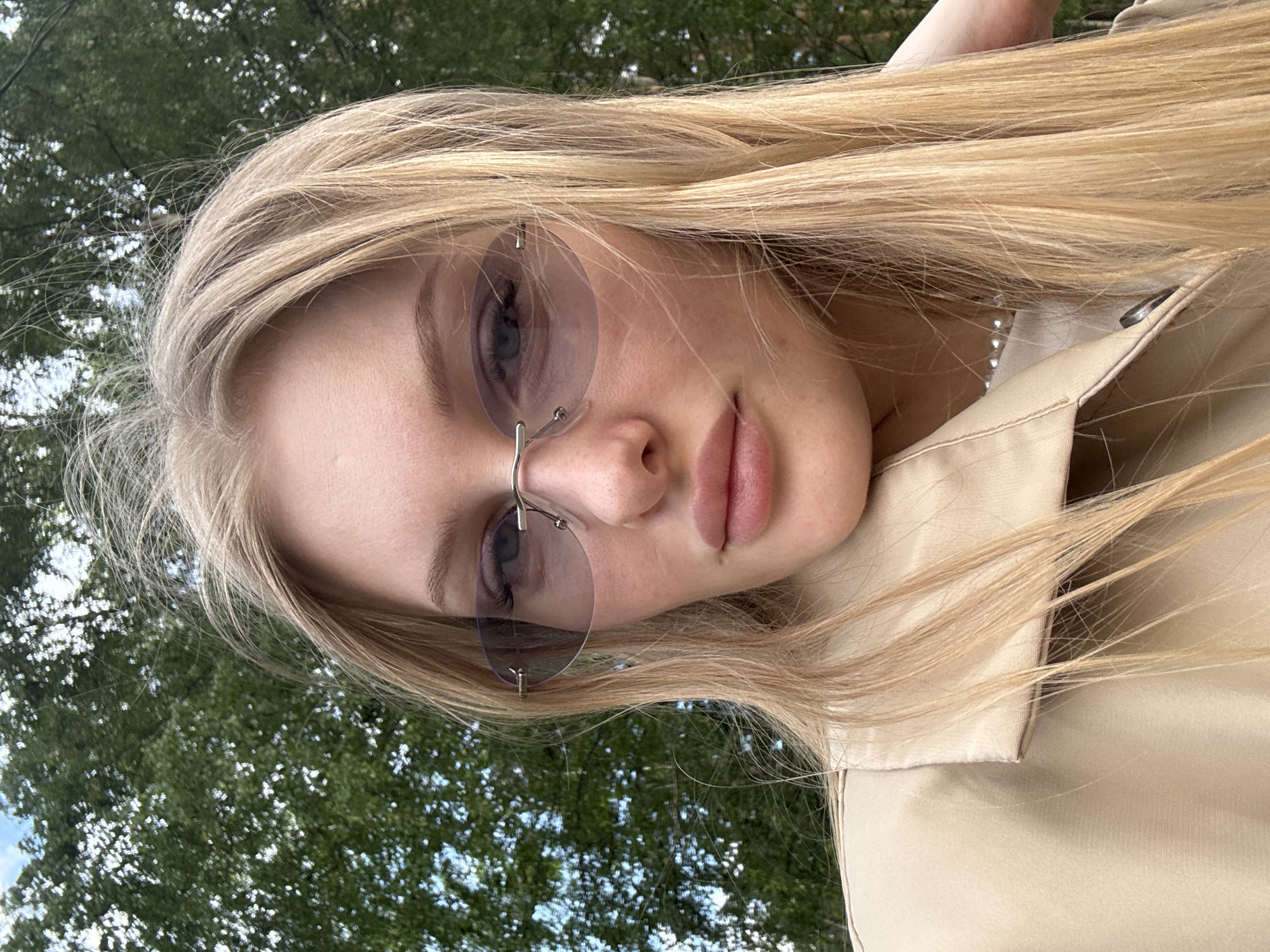 A close-up outdoor portrait features a young woman with long, straight blonde hair, angled diagonally. She is wearing rimless, oval-shaped sunglasses with a light purple tint, which partially obscure her left eye. Her lips are slightly parted in a neutral expression. A pearl necklace is visible under her hair on the right side, along with the collar of a light-colored (beige or tan) jacket or coat. The background consists of vibrant green tree foliage interspersed with patches of bright blue sky, indicating a sunny daytime setting in a natural environment such as a park or garden. The image captures a casual moment or selfie. No text is visible.