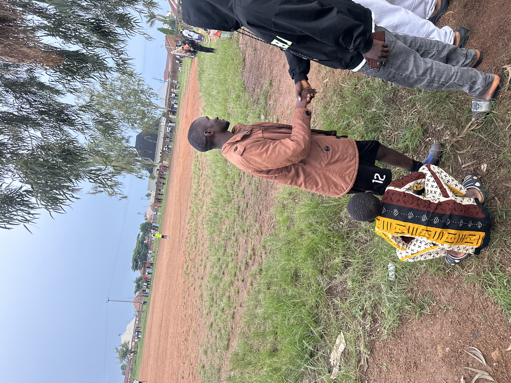 In Du, Nigeria, at a dusty outdoor field with sparse grass, two men are shaking hands. The man on the left, wearing a brown jacket over a dark t-shirt and black shorts with the number '12' on them, faces right. The man on the right wears a black hoodie and grey jeans, and holds a phone in his right hand. To the left of the handshake, a child in a colorful patterned outfit sits on the ground, facing away from the camera. In the background, a crowd of people stands along the edge of the field, and a soccer game appears to be in progress on the reddish-brown dirt surface. Trees and some buildings are visible in the distance under an overcast sky.