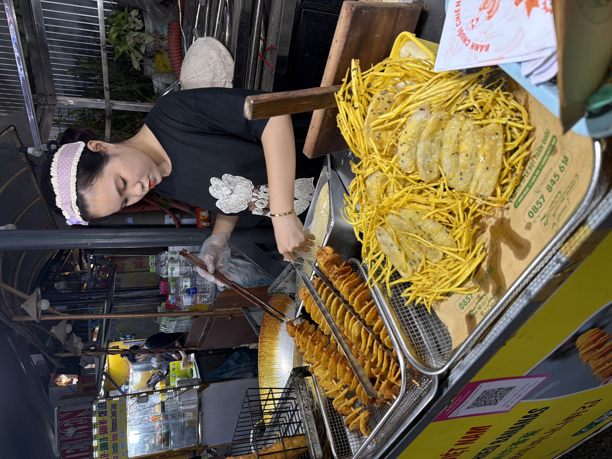 In Đà Nẵng, Vietnam, a street vendor prepares fried bananas at a food stall. The vendor, a young woman with a purple and white polka-dot headband and a black t-shirt, is seen holding chopsticks and tongs. In front of her are two trays of fried food. The tray closest to the viewer contains sliced bananas coated in a yellow batter and topped with black sesame seeds, some of which are entangled with thin, crispy fried strands. Behind this tray, another tray holds spiral-cut fried potatoes or similar root vegetables on skewers. The stall itself is brightly lit with yellow signage, and in the background, there are shelves displaying bottled drinks and other food items. The scene captures a moment of food preparation in an outdoor market setting.