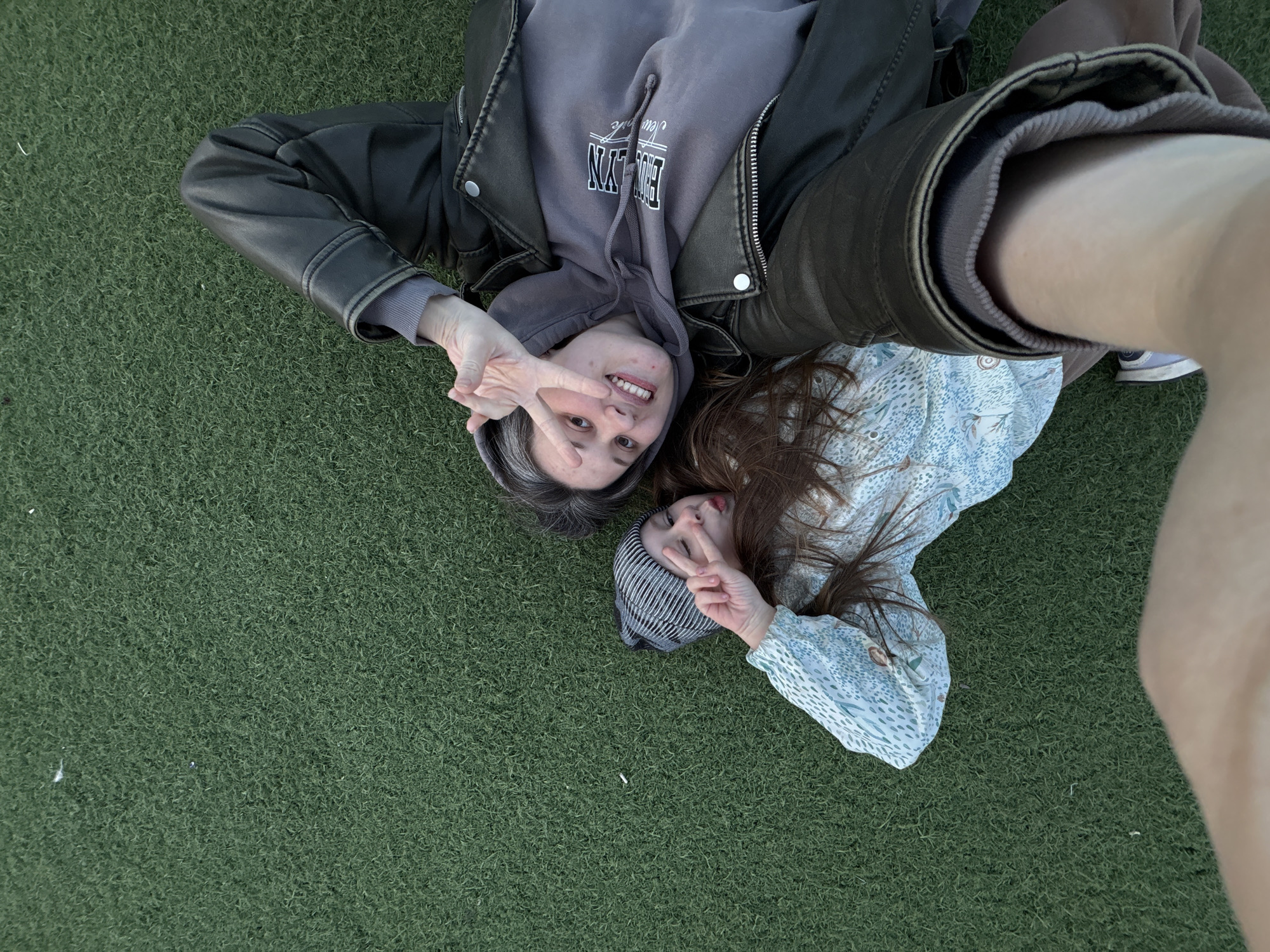 Two people are lying on a patch of green artificial turf. The person on the left is wearing a leather jacket over a gray hooded sweatshirt with white lettering, and is making a peace sign with their fingers. Their hair is graying. The person on the right is wearing a striped beanie and a blue patterned top, and is also making a peace sign. A person's arm and part of their torso is visible on the right edge of the image.
The city of Chaikovsky, Russia, is not discernible in the image.